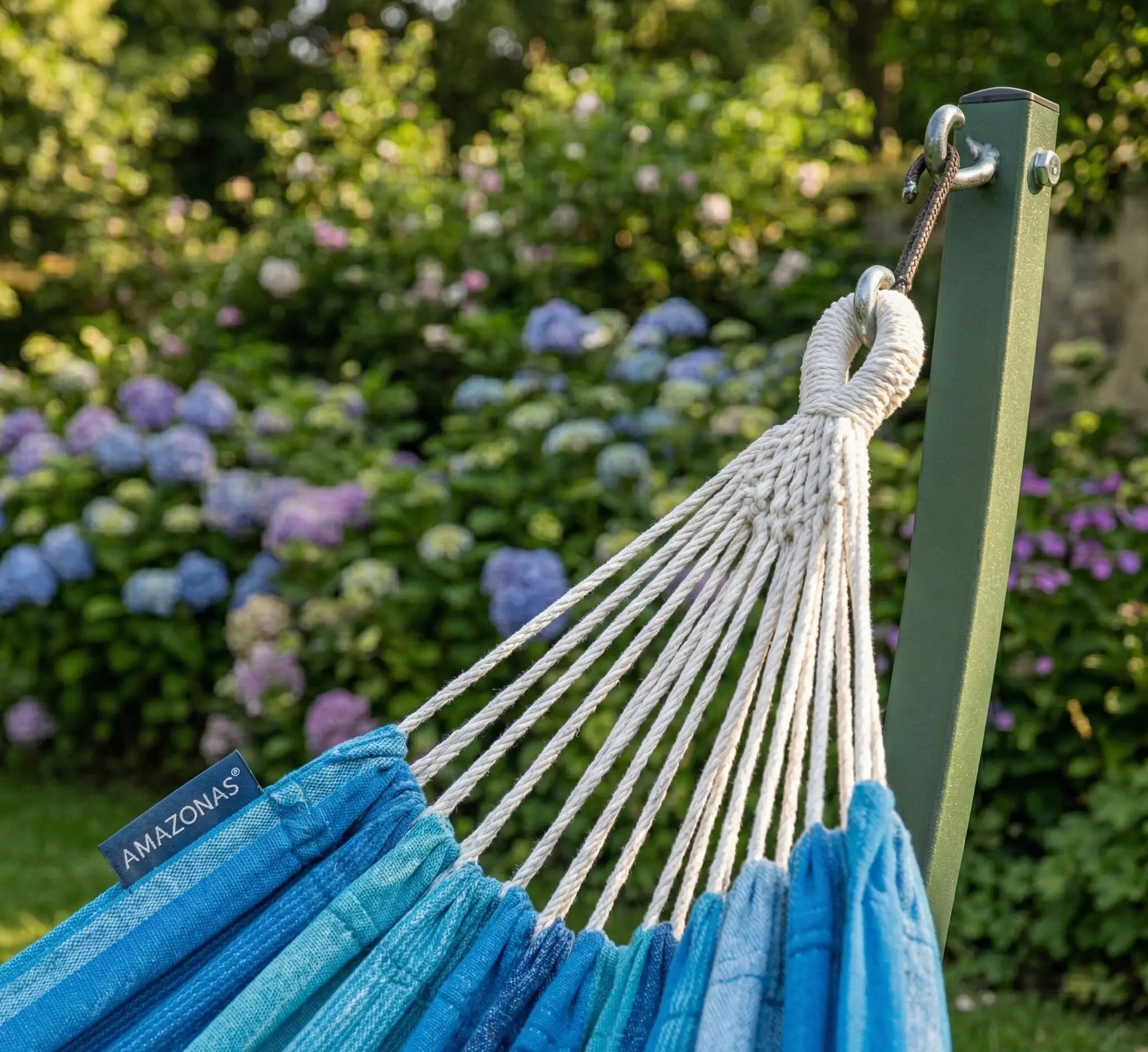 Détail de la boucle de suspension tressée blanche d'un hamac bleu lagon fixée sur un montant en acier vert forêt robuste dans un jardin avec hortensias