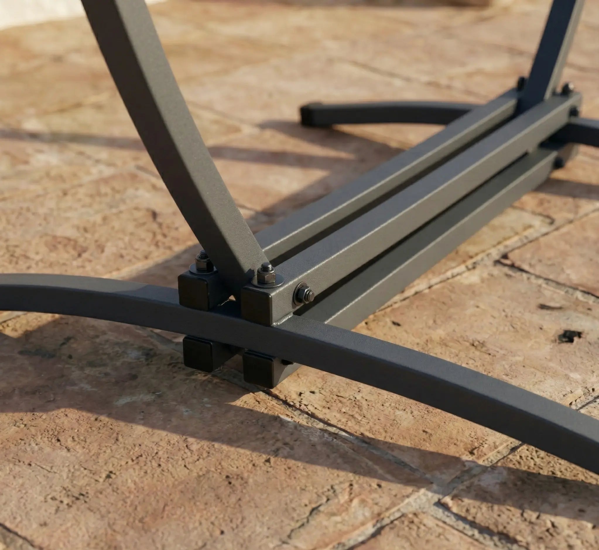 Détail de la base en acier noir d'un support de chaise hamac autoportant, montrant les fixations boulonnées et les pieds incurvés stables posés sur une terrasse en dalles de pierre au soleil