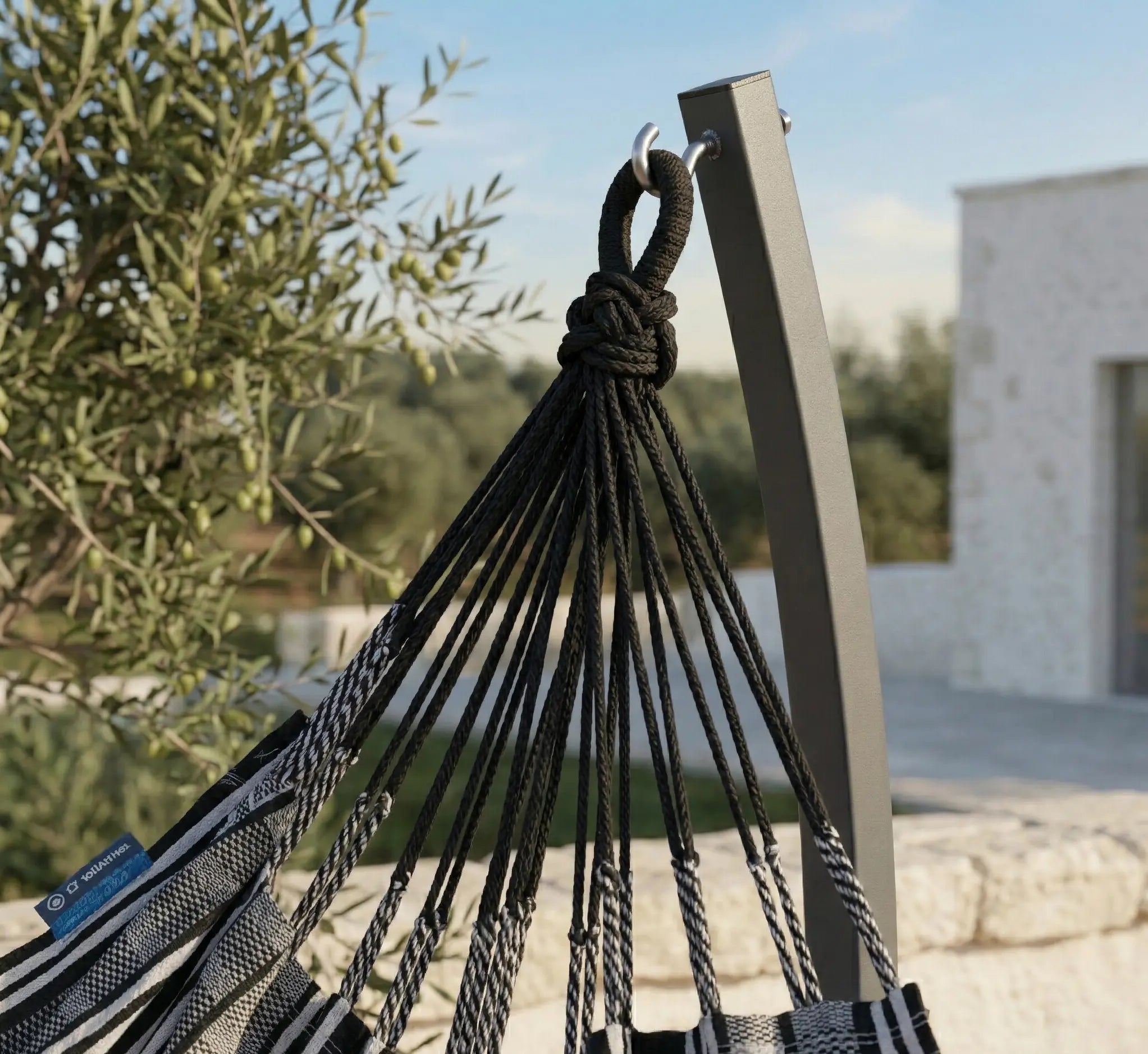 Chaise hamac en toile à rayures bleues et blanches suspendue à un support métallique noir en arc dans un jardin verdoyant et fleuri avec vue sur l'horizon ensoleillé