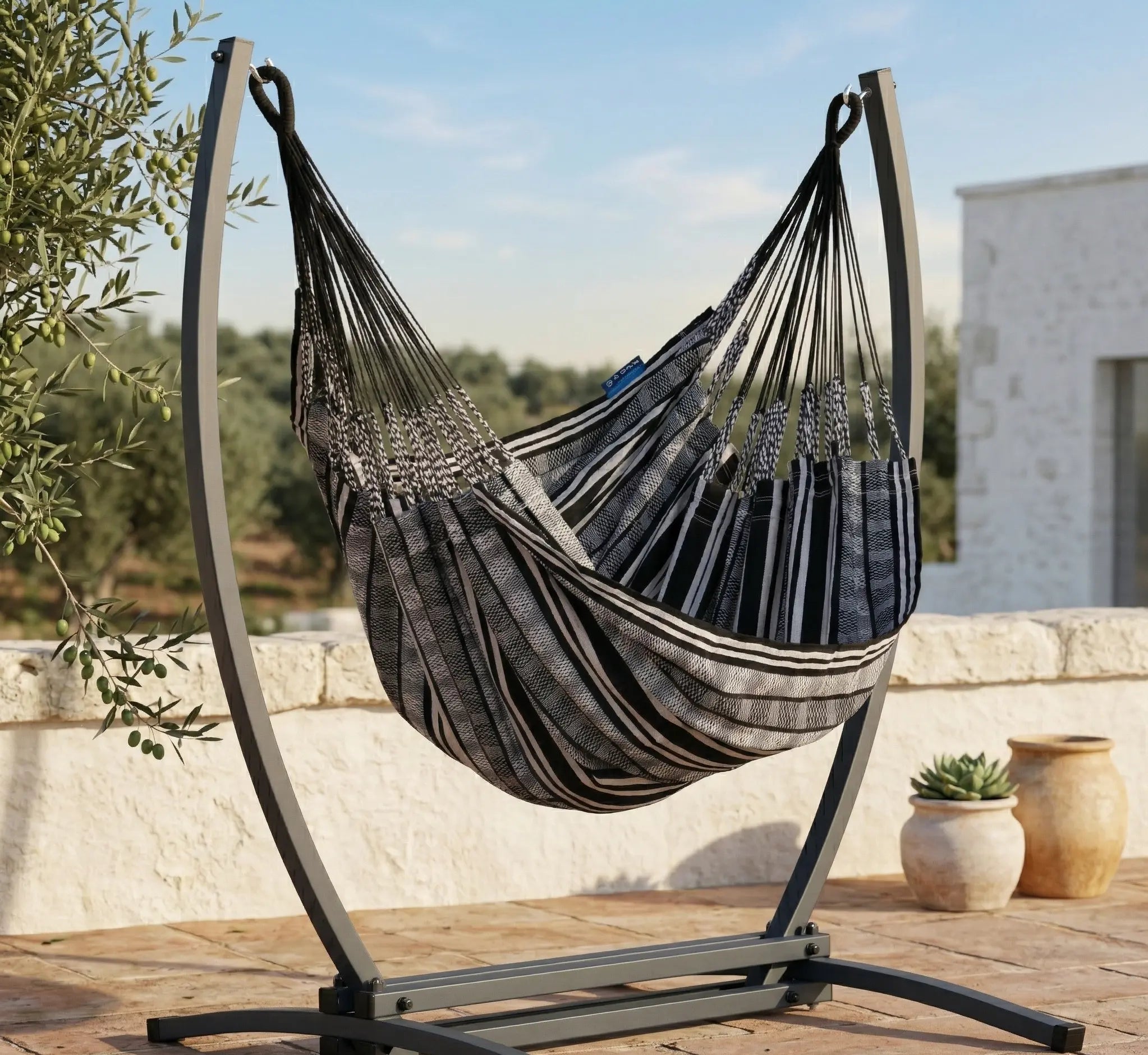 Chaise hamac en toile à rayures noires et grises suspendue à un support métallique noir en arc dans un jardin verdoyant et fleuri avec vue sur l'horizon ensoleillé