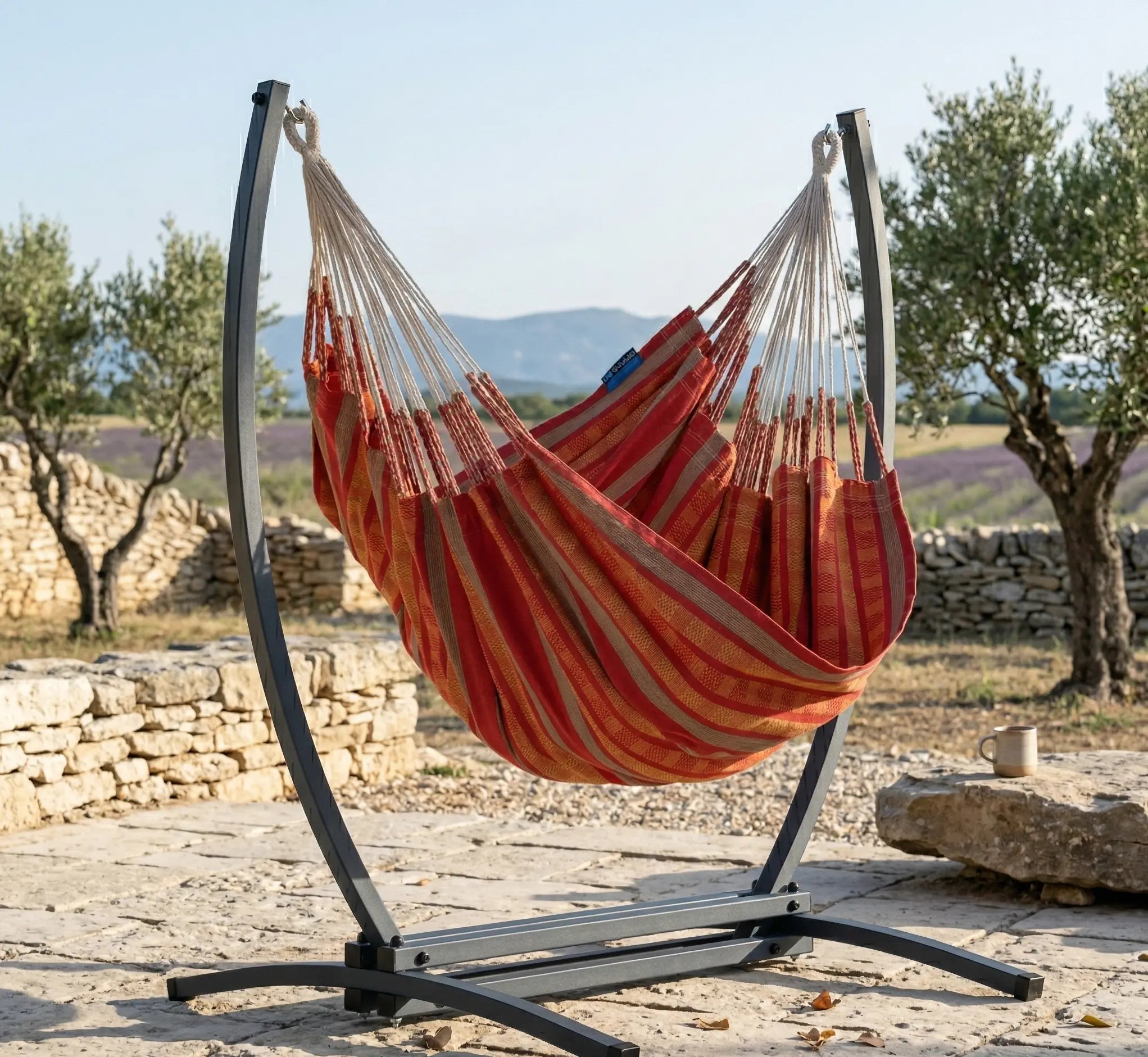 Chaise hamac en toile à rayures rouges et orange suspendue à un support métallique noir en arc dans un jardin verdoyant et fleuri avec vue sur l'horizon ensoleillé