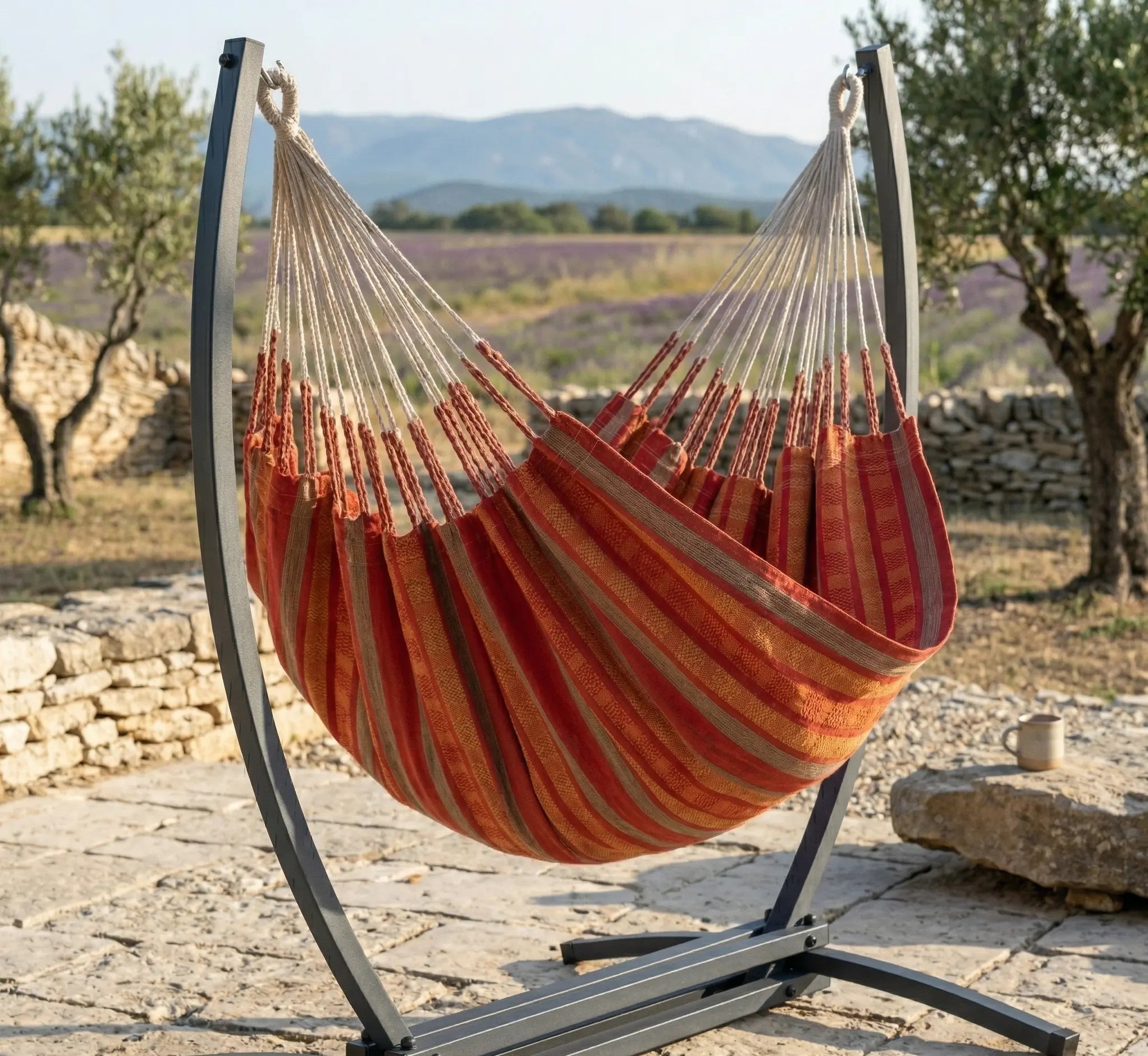 Chaise hamac en toile à rayures rouges et orange suspendue à un support métallique noir en arc dans un jardin verdoyant et fleuri avec vue sur l'horizon ensoleillé