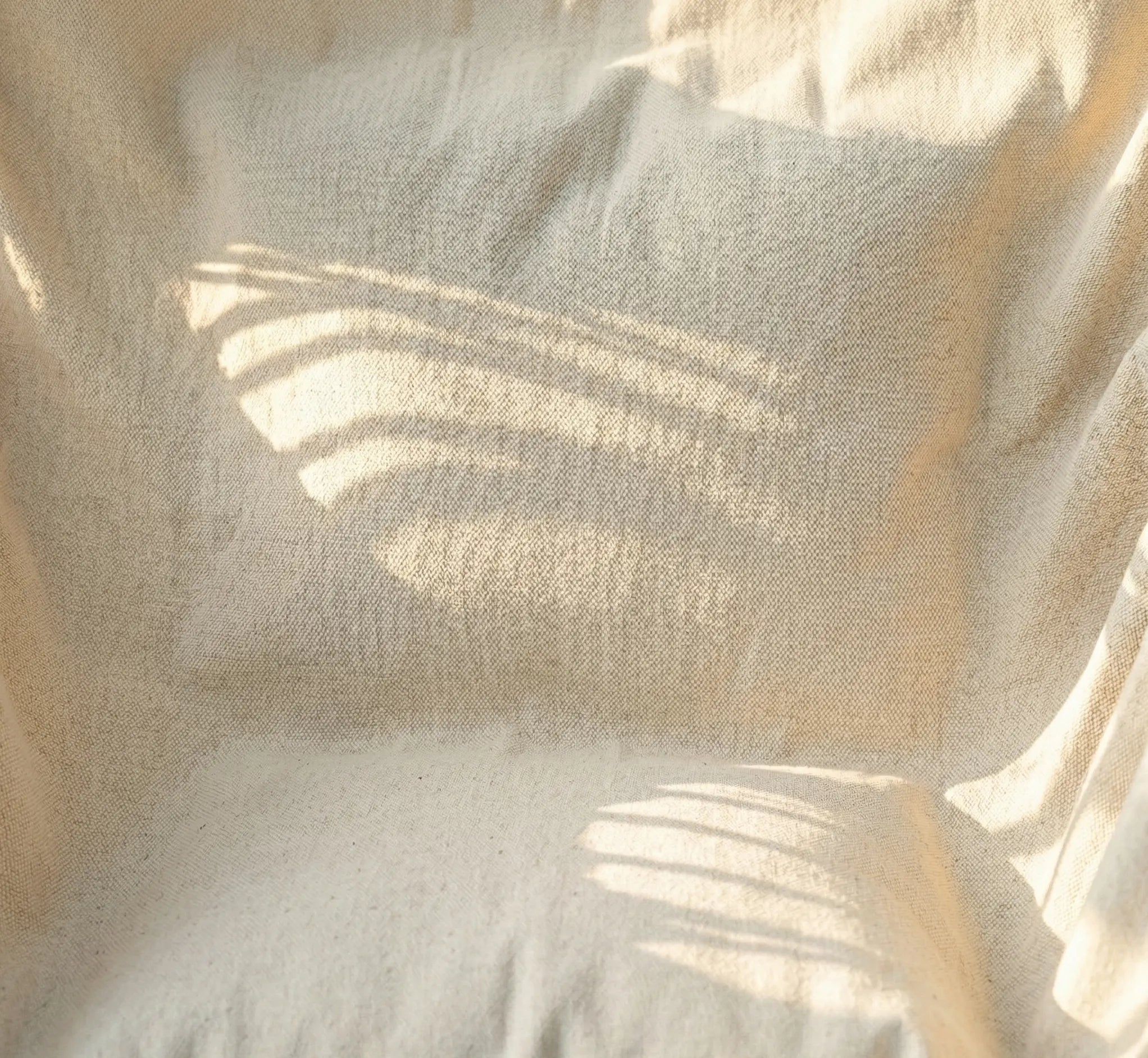 Chaise hamac suspendue en toile beige avec deux coussins moelleux et barre en bois installé dans un jardin verdoyant et fleuri avec mur en pierre en arrière-plan