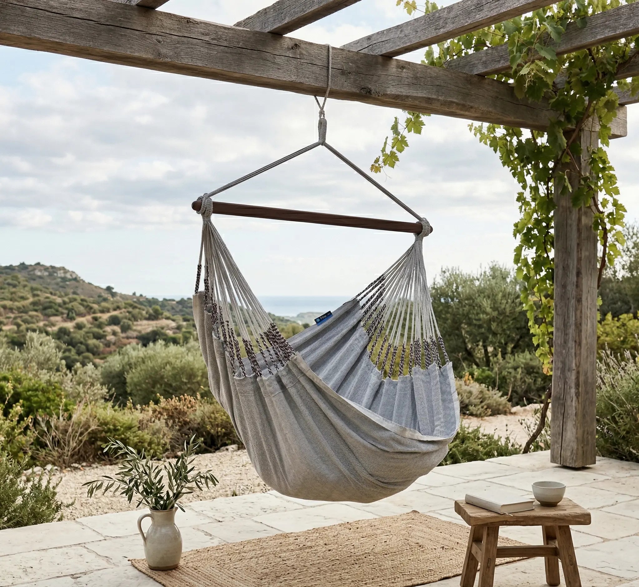 Chaise hamac suspendue en tissu gris avec barre en bois, installée sous une pergola en bois rustique sur une terrasse en pierre avec vue panoramique sur les collines et la mer