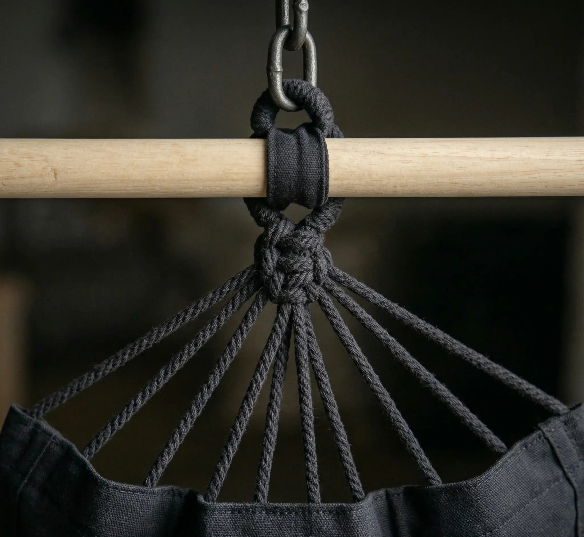 Chaise hamac suspendue en toile noire avec deux coussins moelleux et barre en bois installé dans un jardin verdoyant et ensoleillé avec plantes luxuriantes en arrière-plan