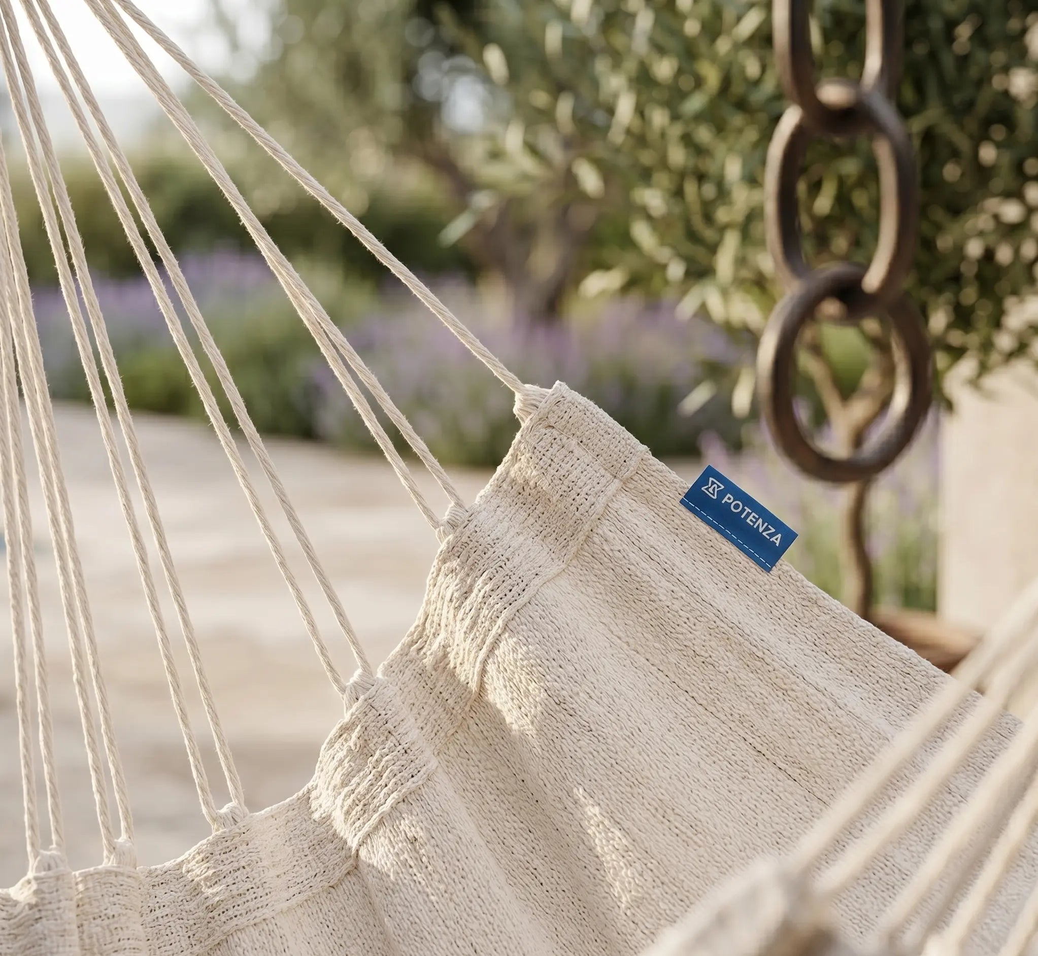 Fauteuil suspendu en toile blanc crème avec barre d'écartement en bois clair, suspendu à une branche d'olivier rustique sur une terrasse en dalles de pierre avec vue sur une vallée verdoyante et ensoleillée.