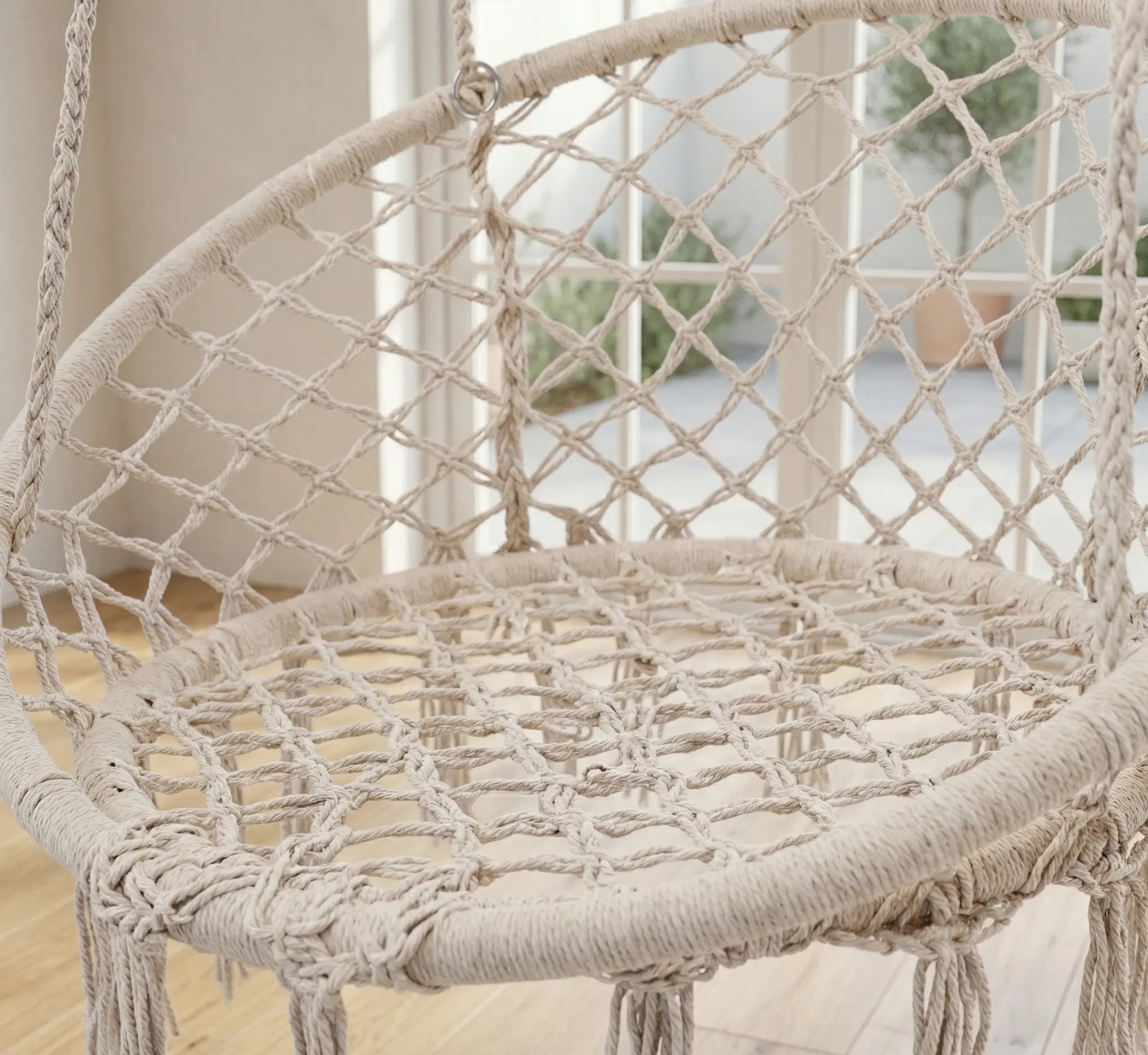 Balancelle ronde en macramé beige avec franges artisanales suspendue sur un balcon ensoleillé face à la mer avec sol en dalles de pierre et plantes vertes
