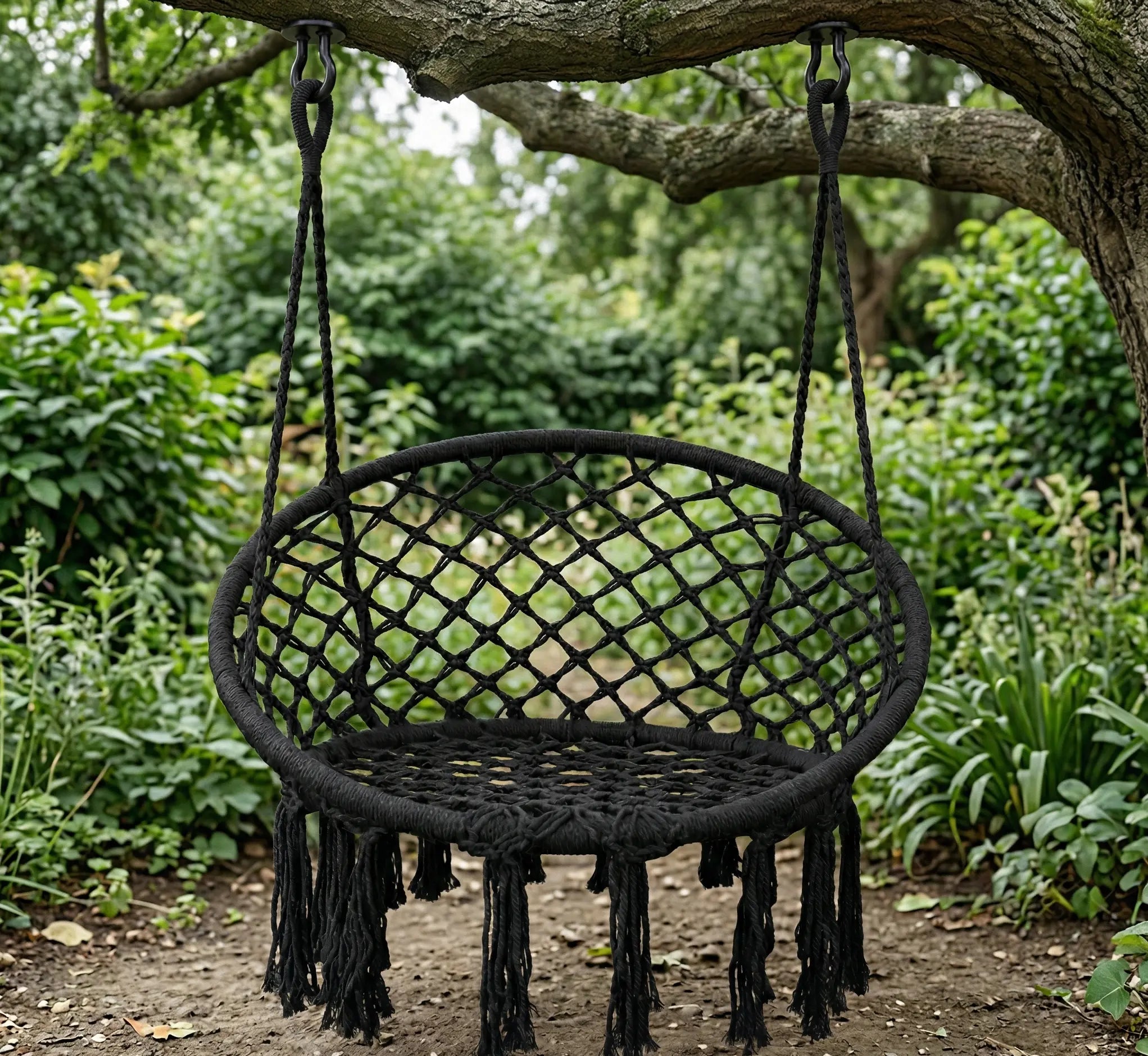 Fauteuil suspendu rond en macramé noir avec franges, accroché à une branche d'arbre dans un jardin verdoyant, créant un coin détente élégant et ombragé