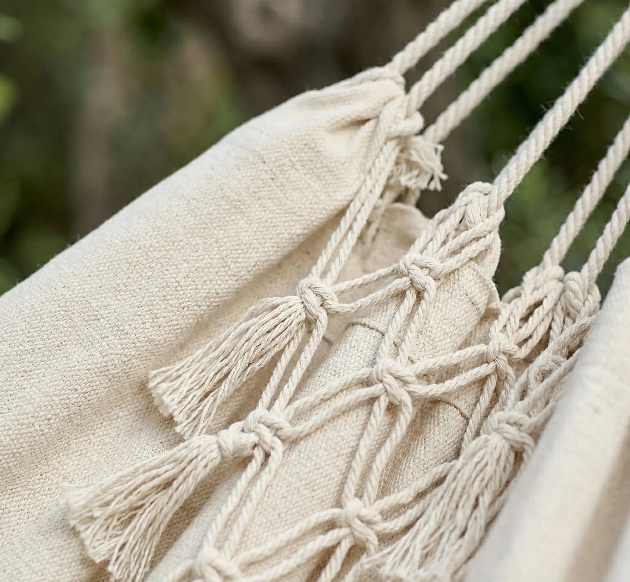 Ambiance bohème chic avec un grand hamac brésilien en coton écru orné de franges en macramé, suspendu entre deux arbres dans un jardin verdoyant et ensoleillé.