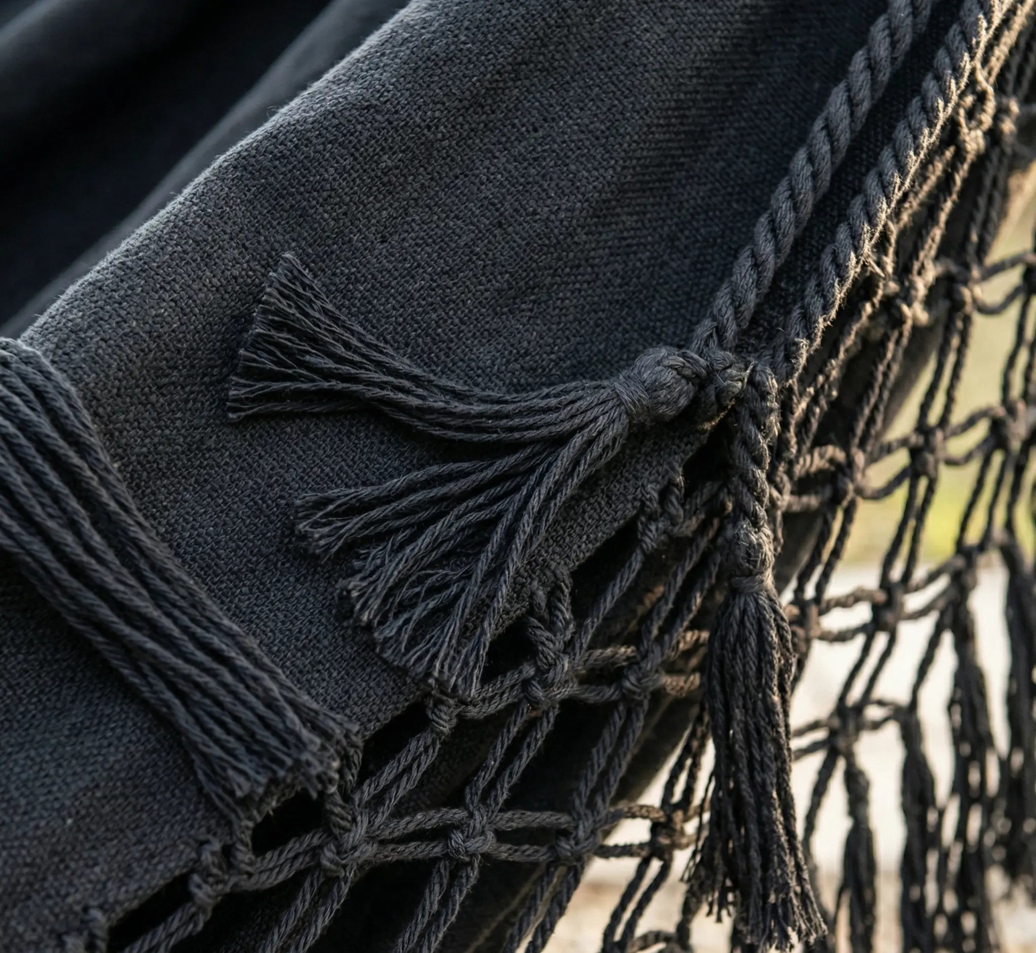 Ambiance bohème chic avec un grand hamac brésilien en coton noir intense orné de franges en macramé, suspendu entre deux arbres dans un jardin verdoyant et ensoleillé.