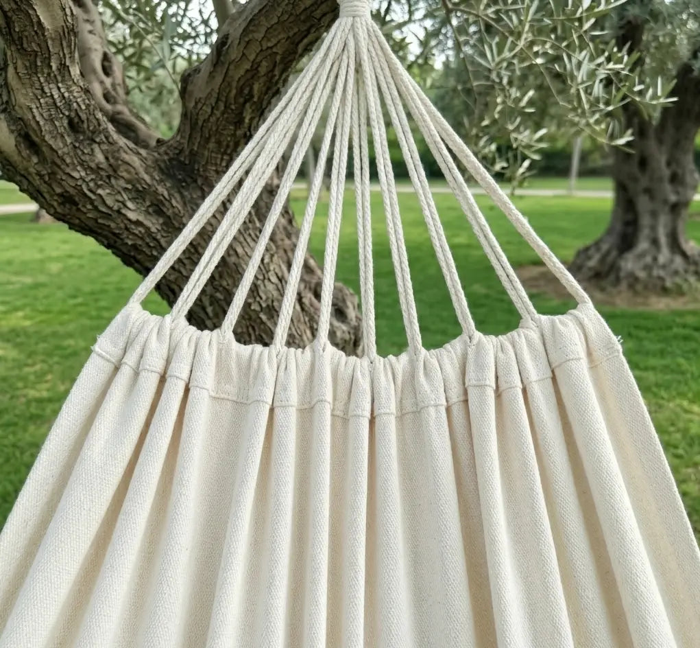 Ambiance bohème avec un grand hamac brésilien en coton écru naturel suspendu entre deux arbres robustes dans un grand jardin paysager.