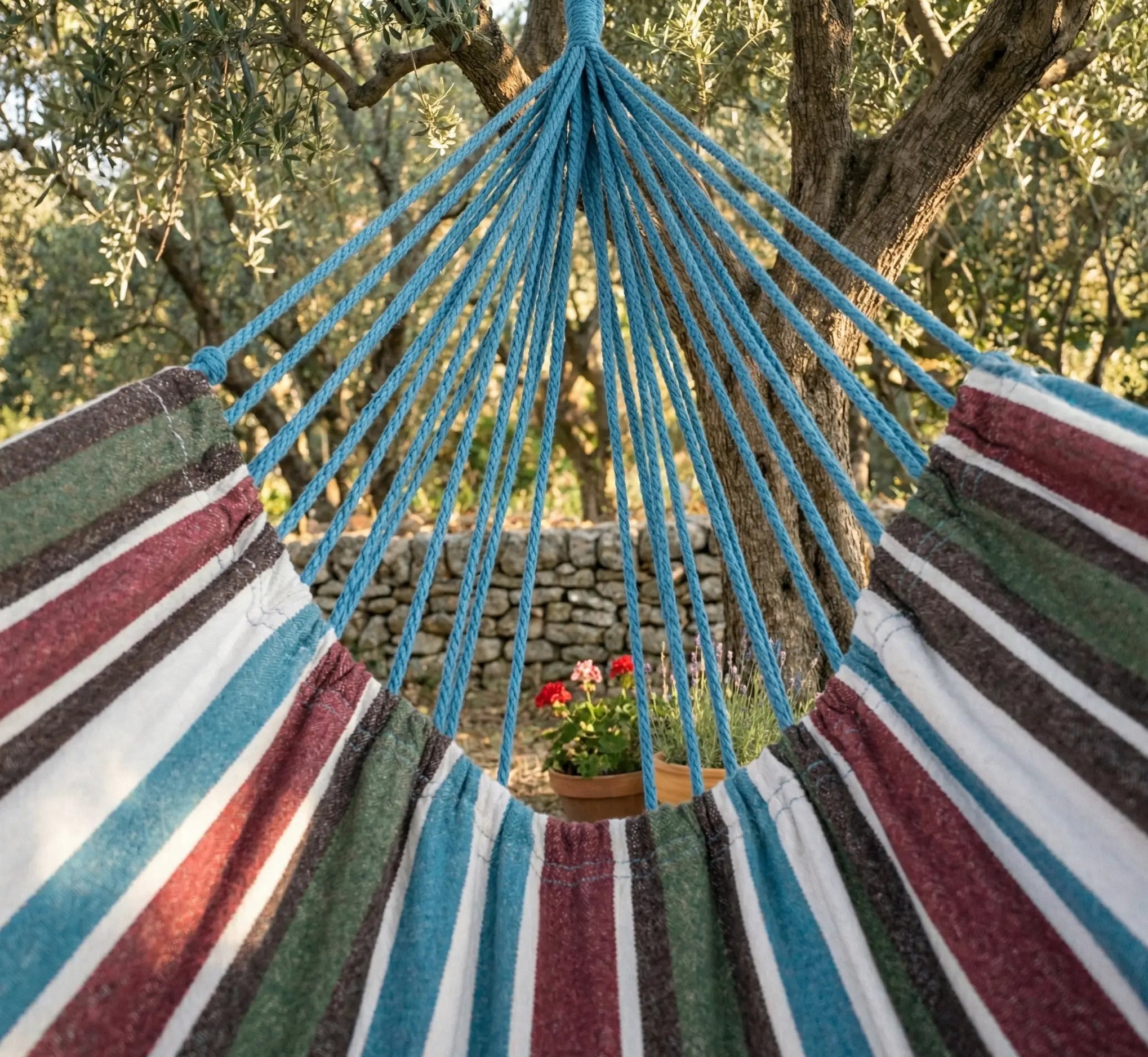 Vue d'ensemble d'un grand hamac brésilien en coton rayé bordeaux bleu et vert, avec des franges et pompons décoratifs, suspendu entre deux arbres dans un jardin verdoyant et ombragé.