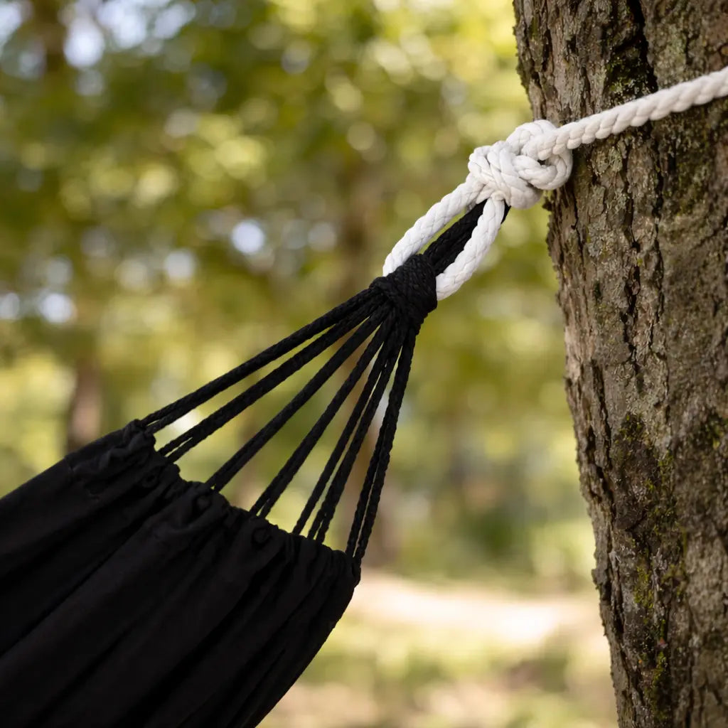 Ambiance moderne avec un grand hamac brésilien en coton noir suspendu entre deux arbres robustes dans un grand jardin verdoyant.