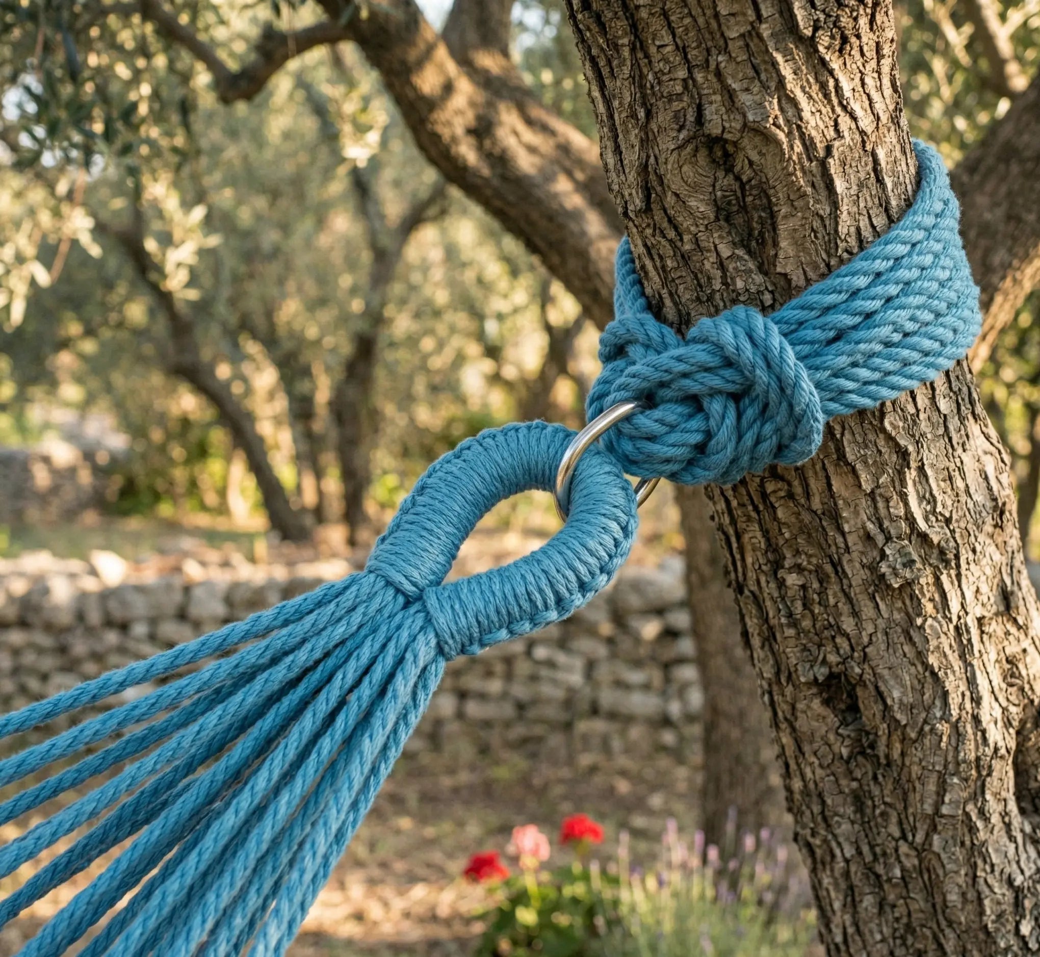 Ambiance détendue avec un grand hamac brésilien en coton à rayures verticales bordeaux vert bleu et blanc suspendu entre deux troncs d'arbres robustes dans un jardin verdoyant et paysager.