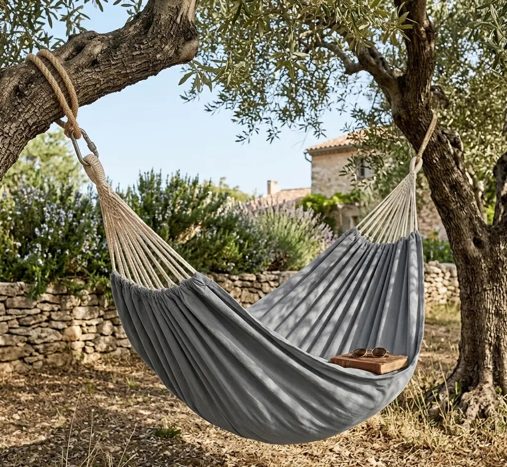 Mise en situation d'un hamac brésilien gris anthracite suspendu entre deux oliviers dans un jardin provençal avec un livre et des lunettes de soleil.