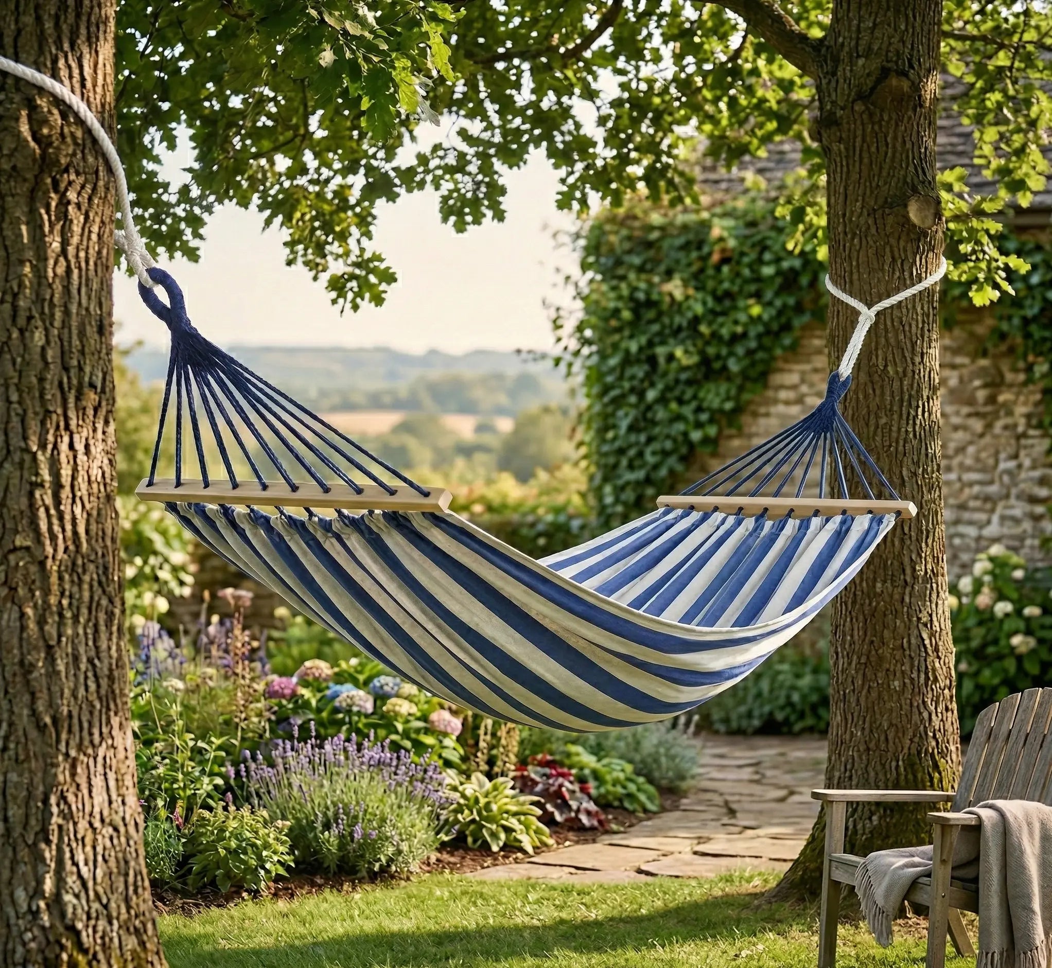 Vue d'un jardin champêtre avec un hamac à rayures bleu et blanc suspendu entre deux arbres, entouré de lavande et d'hortensias, avec une chaise de jardin en bois à droite.