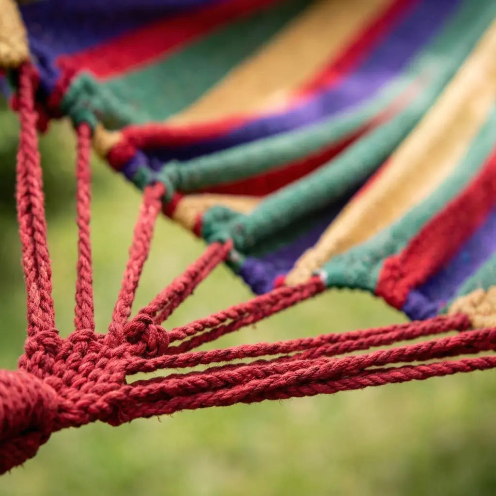 Hamac de jardin multicolore avec barres blanches suspendu entre un arbre et un poteau en bois dans un jardin ensoleillé.