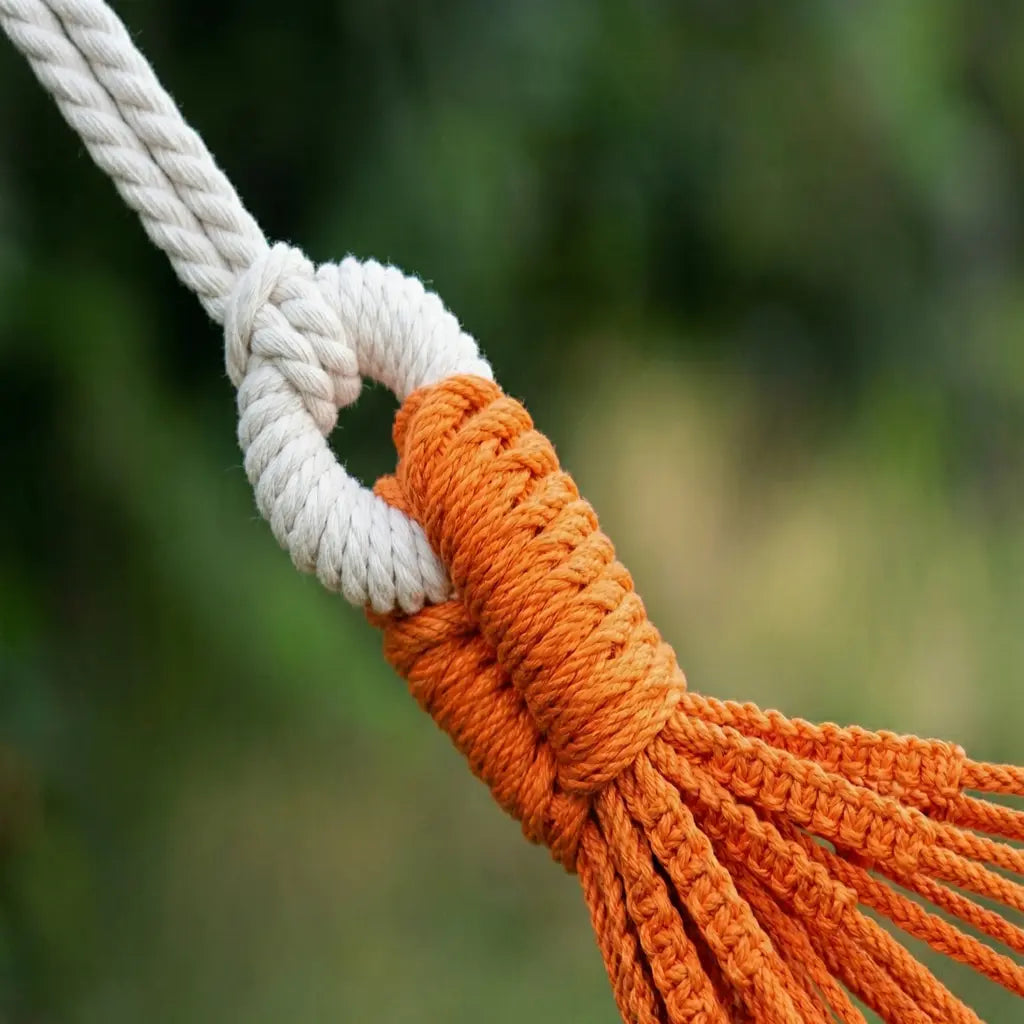 Mise en situation d'un hamac à barres blanches aux rayures orange jaune vert et marron suspendu entre un tronc d'arbre et un support en bois dans un grand jardin ensoleillé.