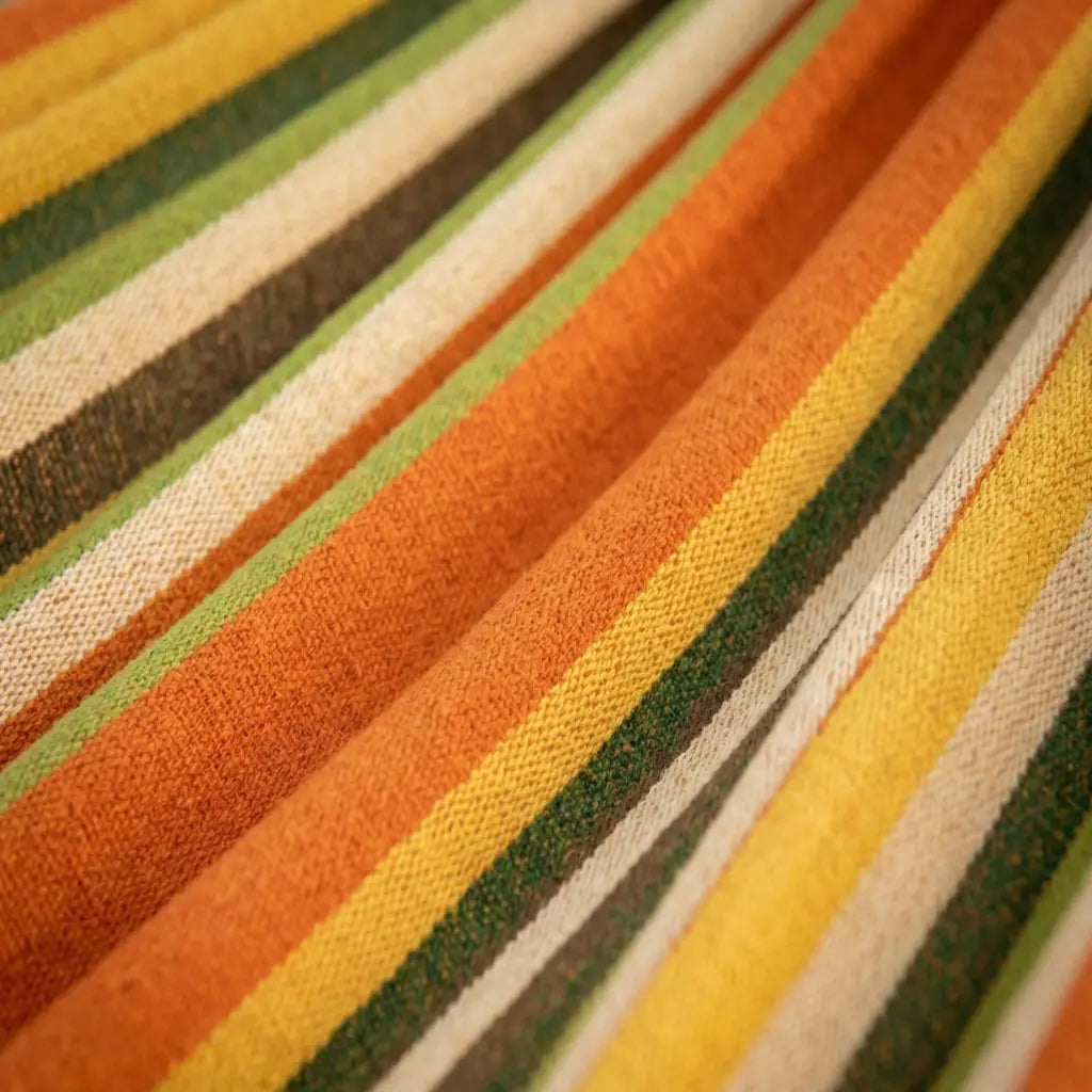 Mise en situation d'un hamac à barres blanches aux rayures orange jaune vert et marron suspendu entre deux arbres dans un grand jardin ensoleillé.