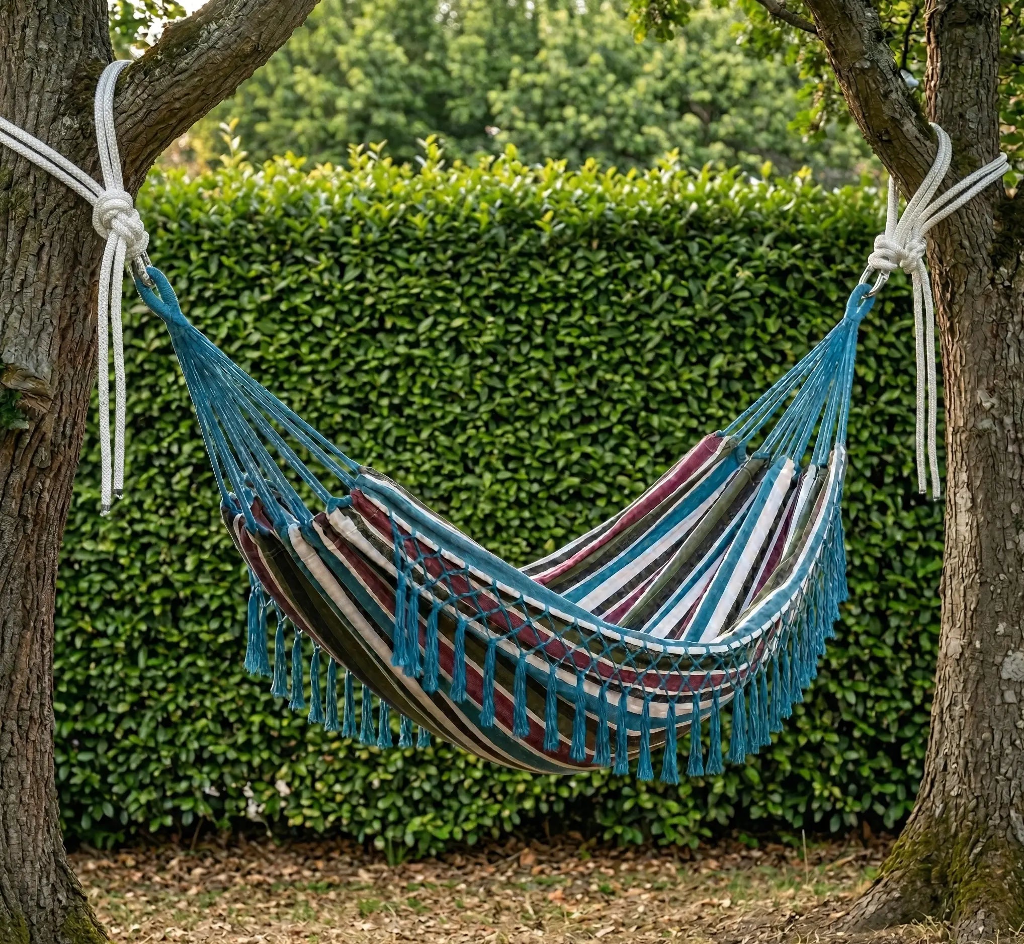 Ambiance détendue avec un grand hamac brésilien en coton à rayures verticales bordeaux vert bleu et blanc suspendu entre deux troncs d'arbres dans un jardin verdoyant.