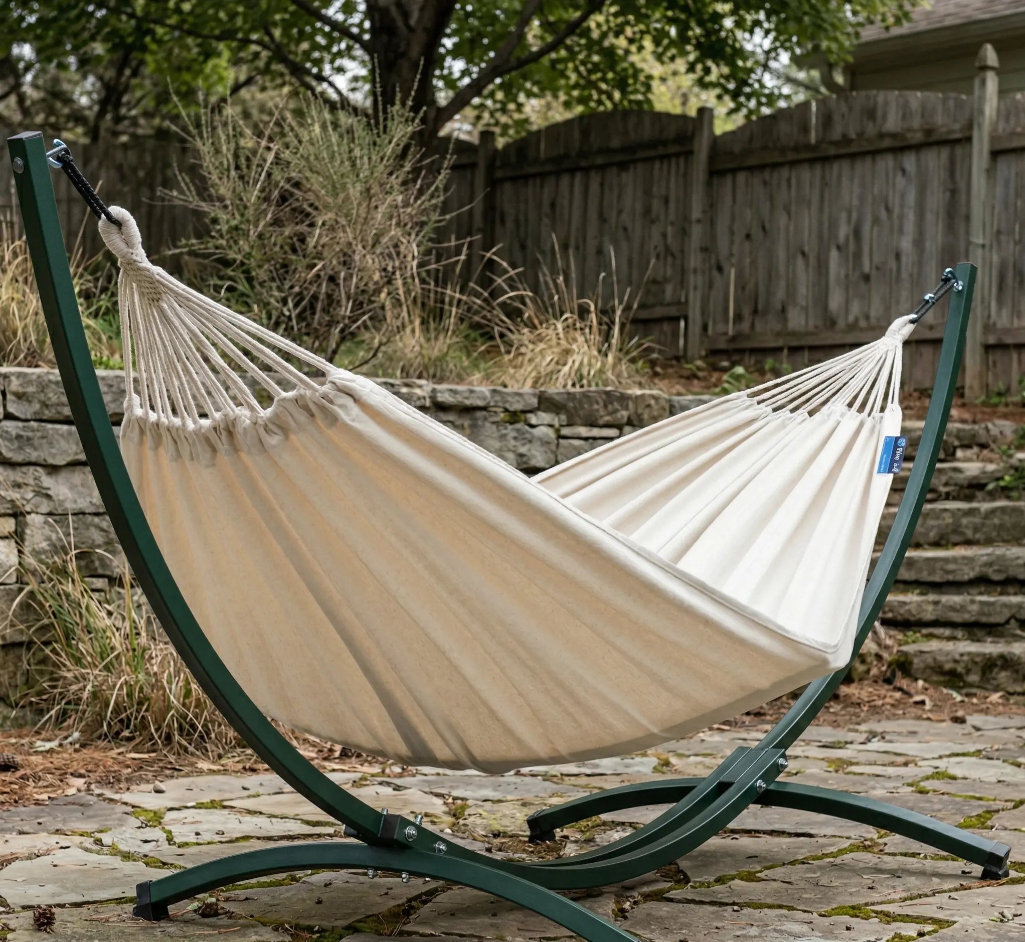 Hamac autoportant avec support courbé en acier vert forêt installé sur une terrasse en pierre, doté d'une toile en coton blanc crème haute densité dans un jardin paysager