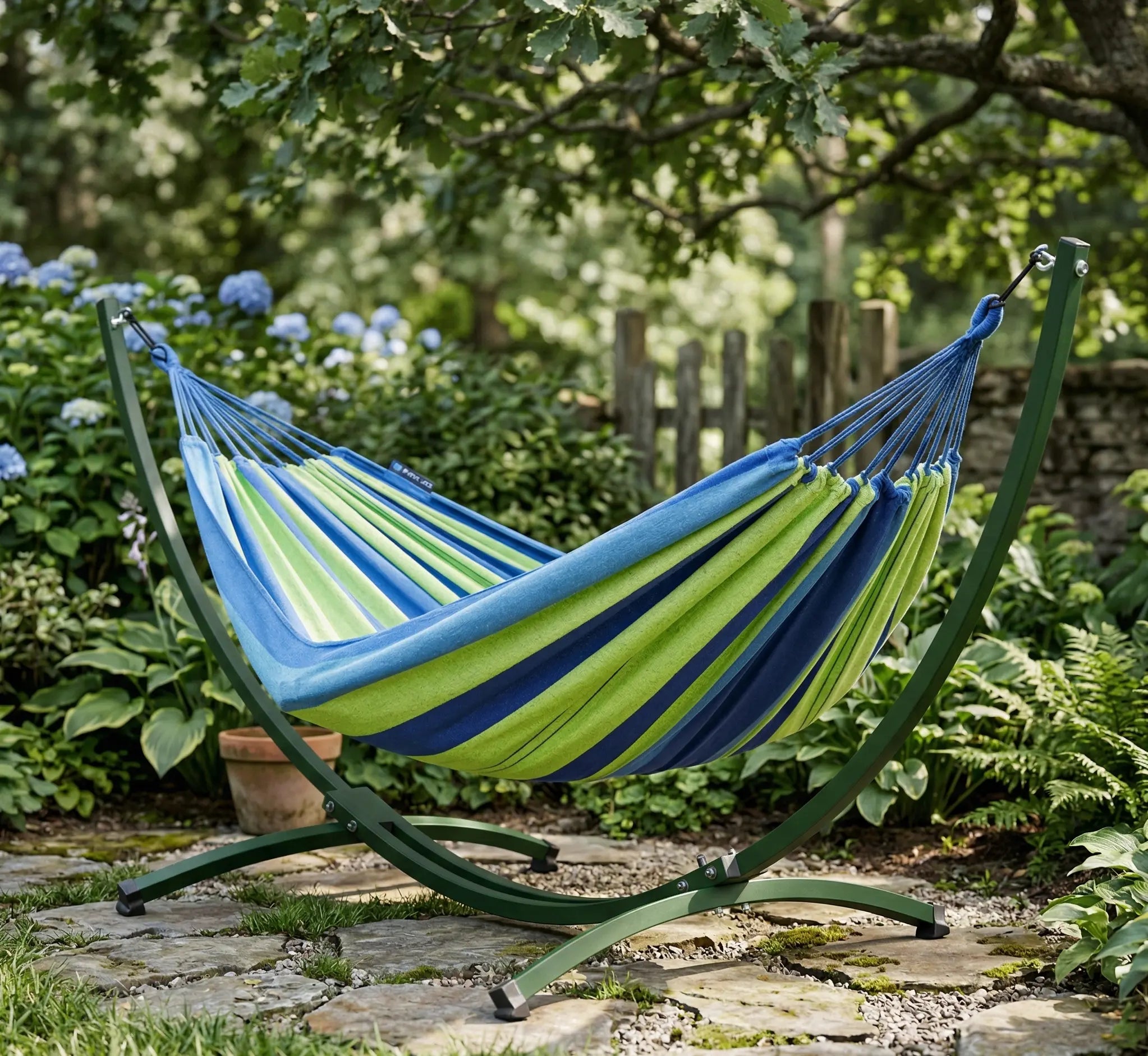 Hamac autoportant avec support en arc acier vert forêt et toile rayée bleu et vert citron installé sur une terrasse en dalles de pierre dans un jardin ombragé