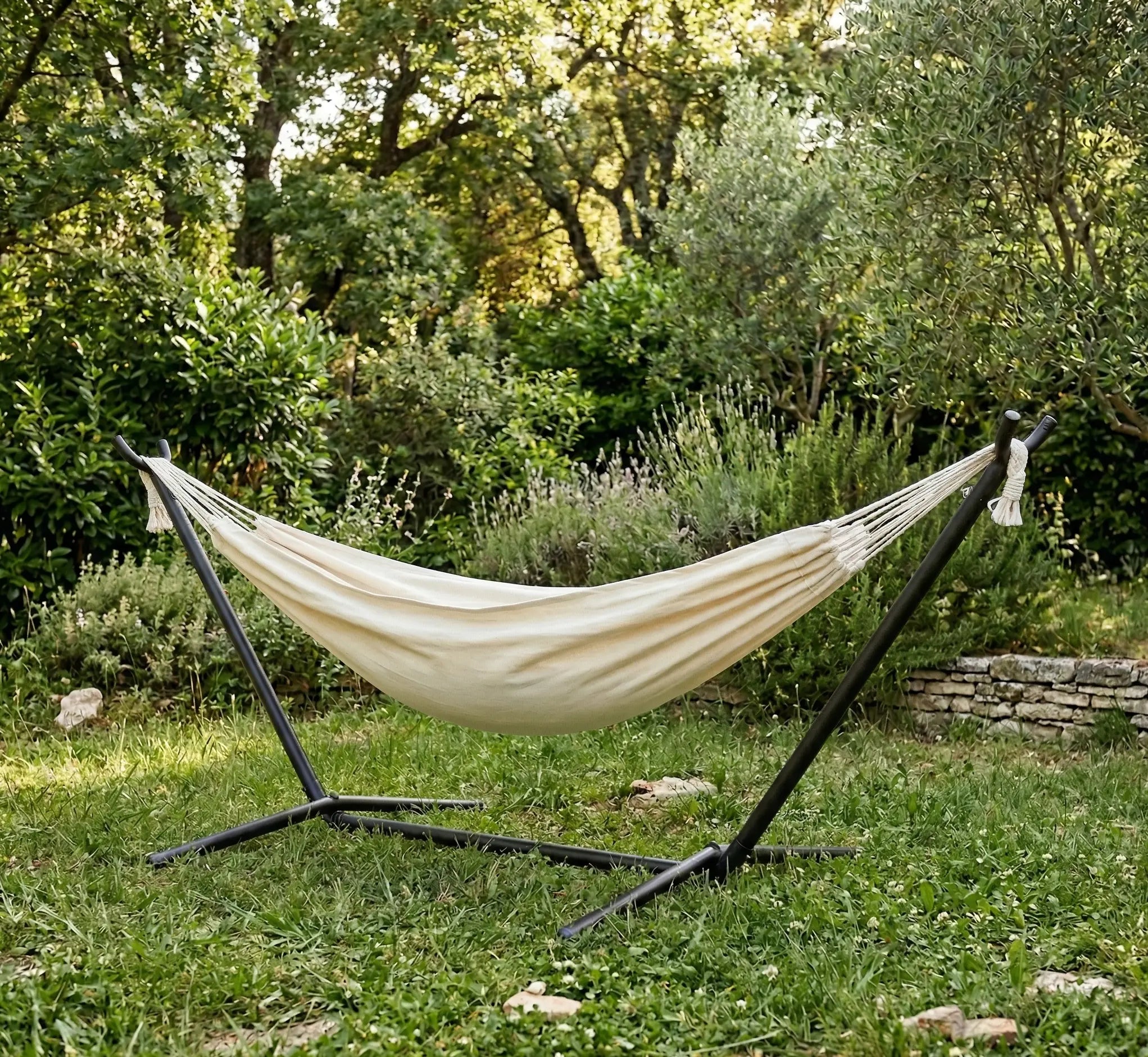 Hamac autoportant avec support en métal noir installé dans un jardin arboré, doté d'une toile en coton blanc crème uni et de cordages tressés de haute qualité