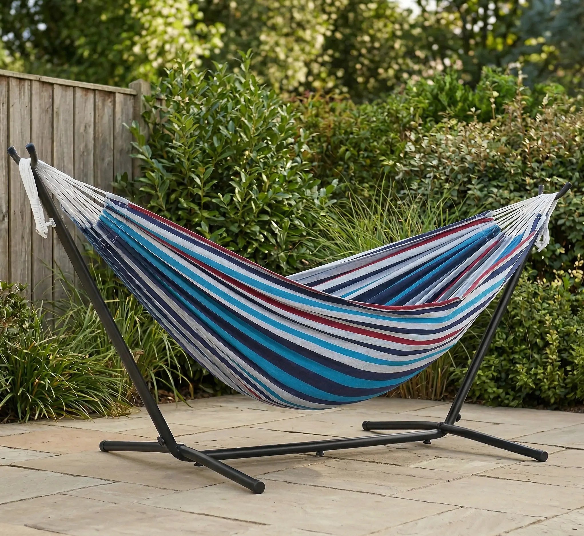 Hamac autoportant haut de gamme installé sur une terrasse en pierre naturelle dans un jardin paysager avec une toile rayée aux tons bleus et bordeaux