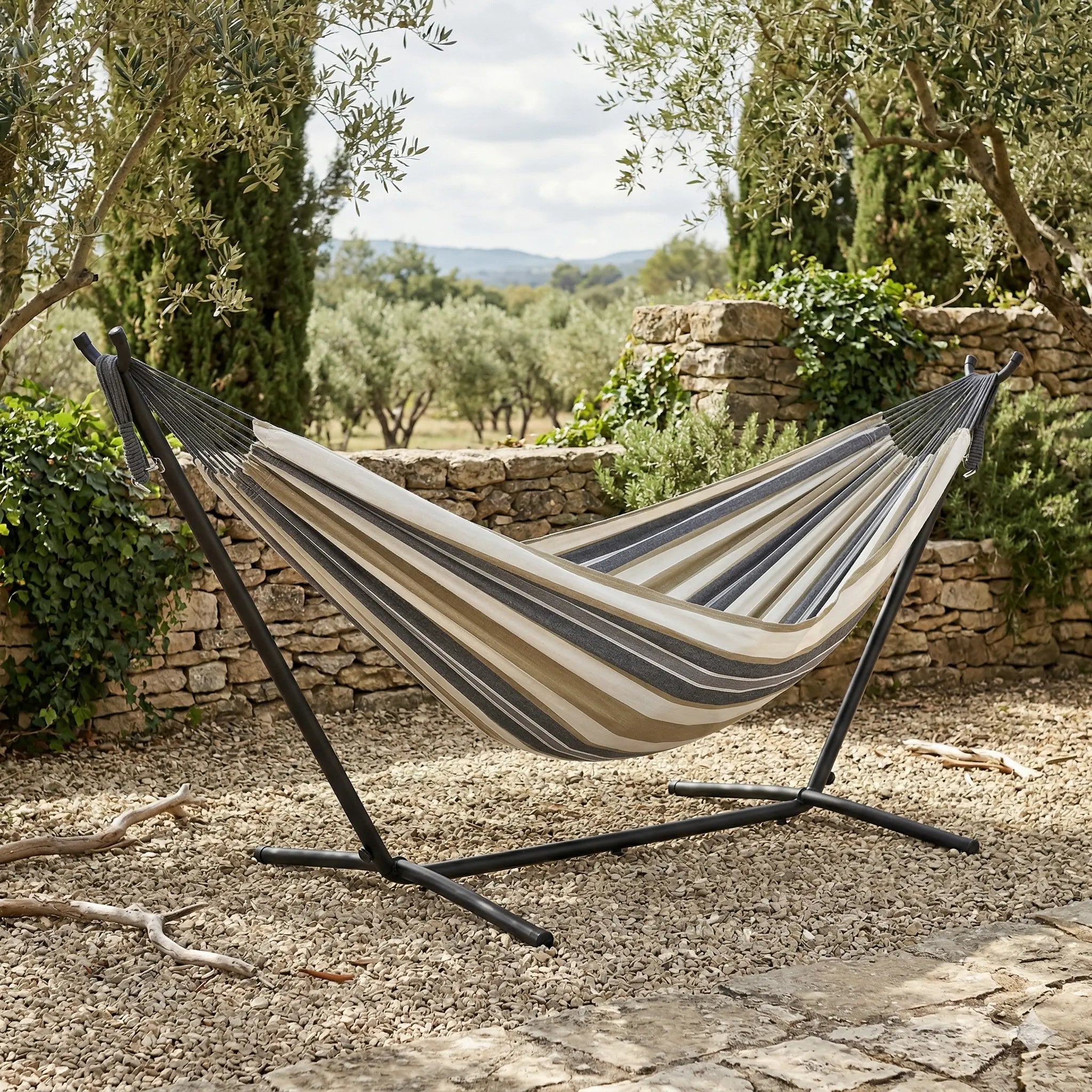 Hamac autoportant avec support en acier noir installé dans un jardin méditerranéen avec muret en pierre et oliviers montrant une toile en coton aux rayures beige et gris anthracite
