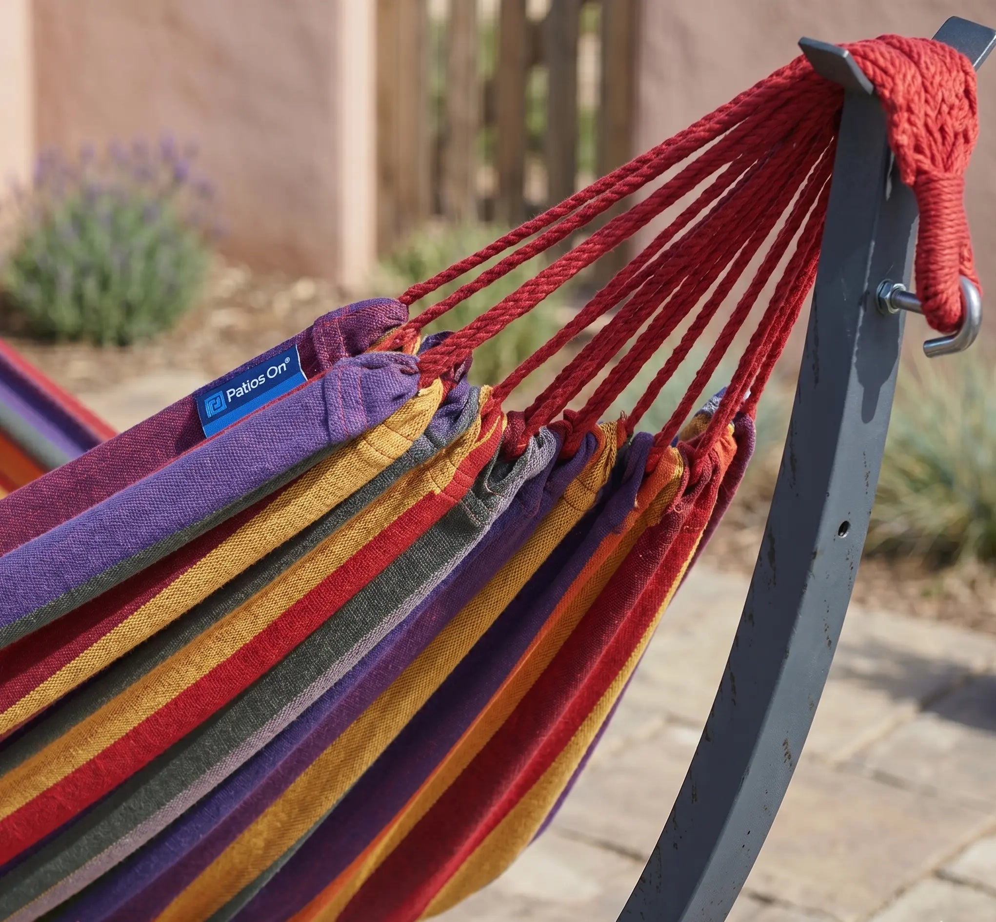 Détail des cordages de suspension rouges fixés à un crochet en acier sur un support gris, montrant une toile de hamac épaisse à rayures violettes, orange et jaunes en extérieur.