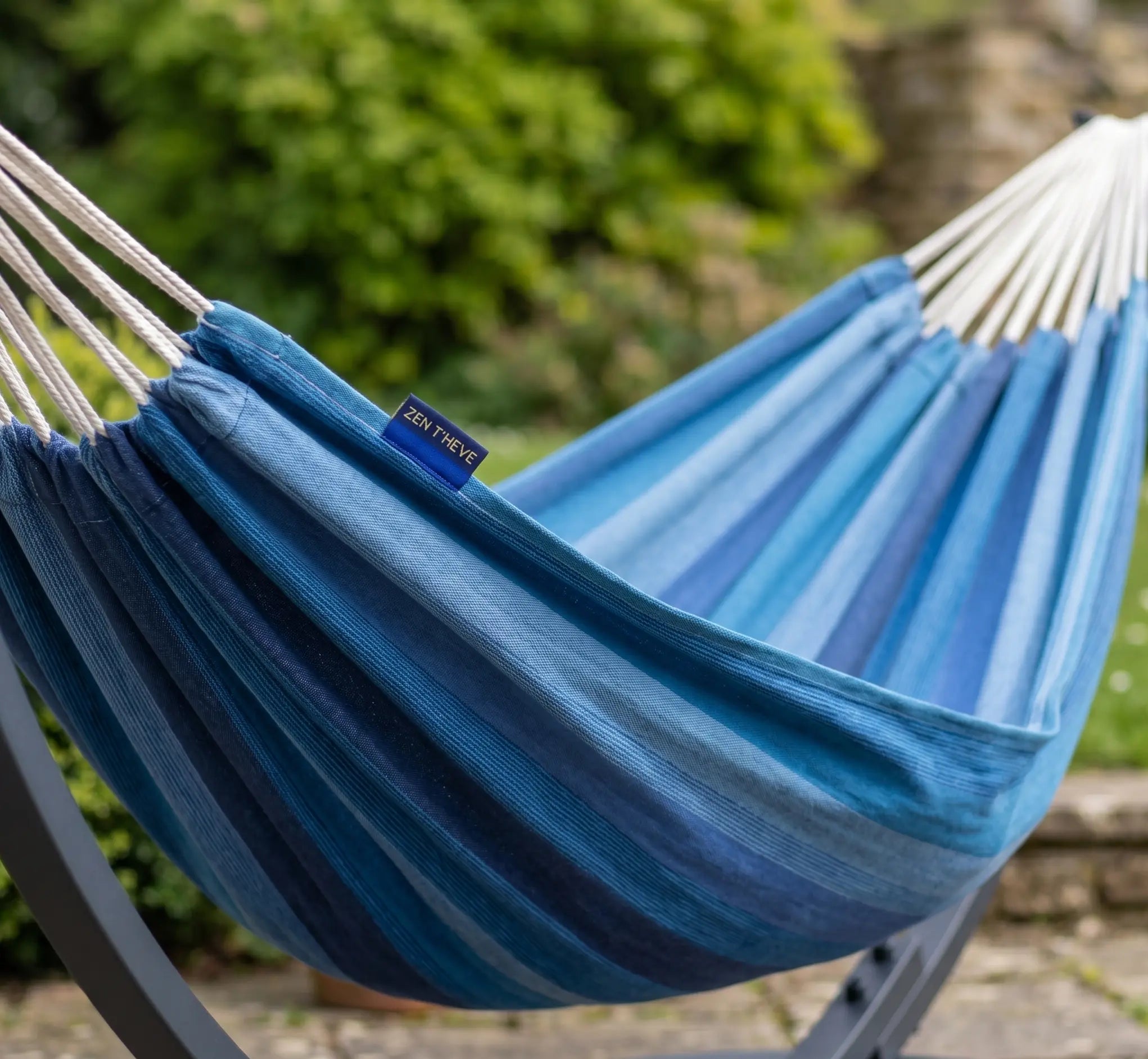 Détail rapproché d'un hamac en toile de coton à rayures bleues dégradées, suspendu à une structure métallique dans un jardin avec un arrière-plan de verdure flou.