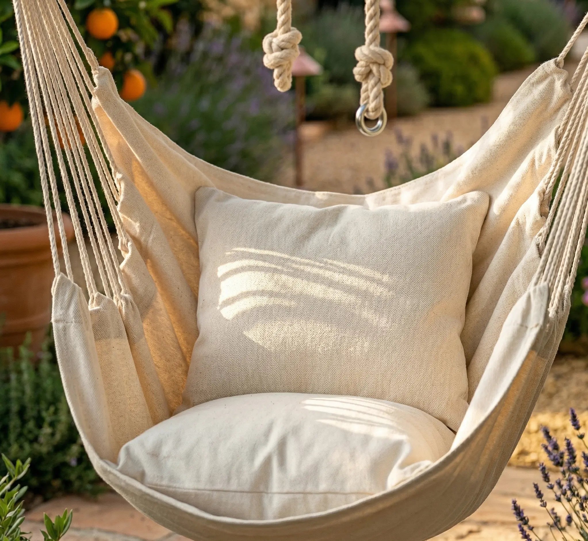 Gros plan sur l'assise d'une chaise hamac beige avec deux coussins moelleux installée dans un jardin ensoleillé, montrant la texture du coton naturel et les jeux d'ombre
