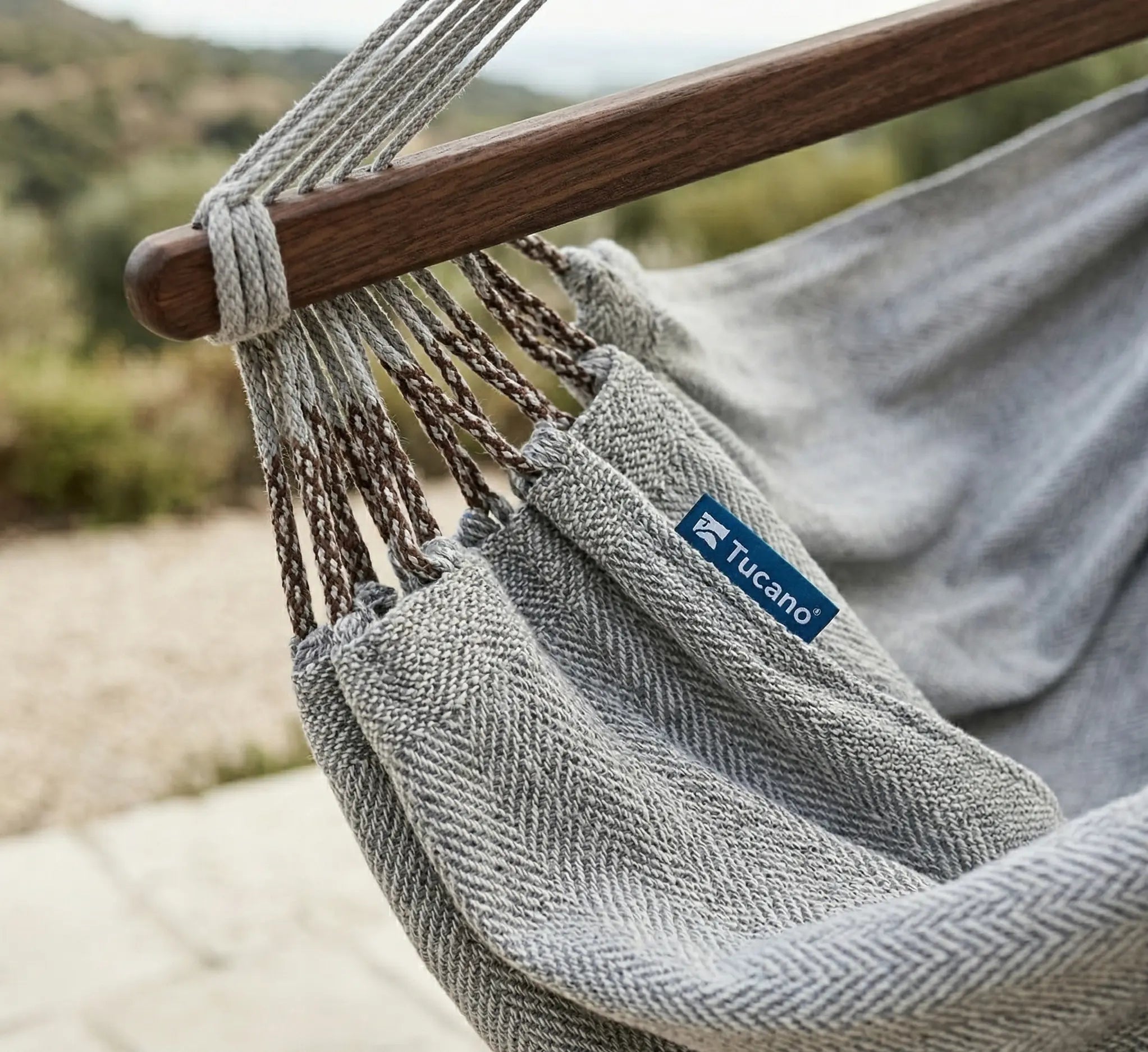 Détail rapproché d'une chaise hamac grise en coton avec tissage à motifs chevrons, montrant la barre d'écartement en bois foncé et les finitions soignées des cordages de suspension