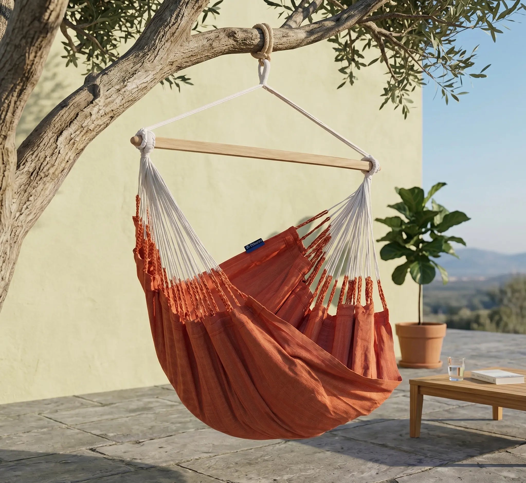 Chaise hamac en toile terracotta suspendue à une branche d'olivier par une corde robuste, avec barre d'écartement en bois, sur une terrasse en pierre ensoleillée avec vue sur la nature.