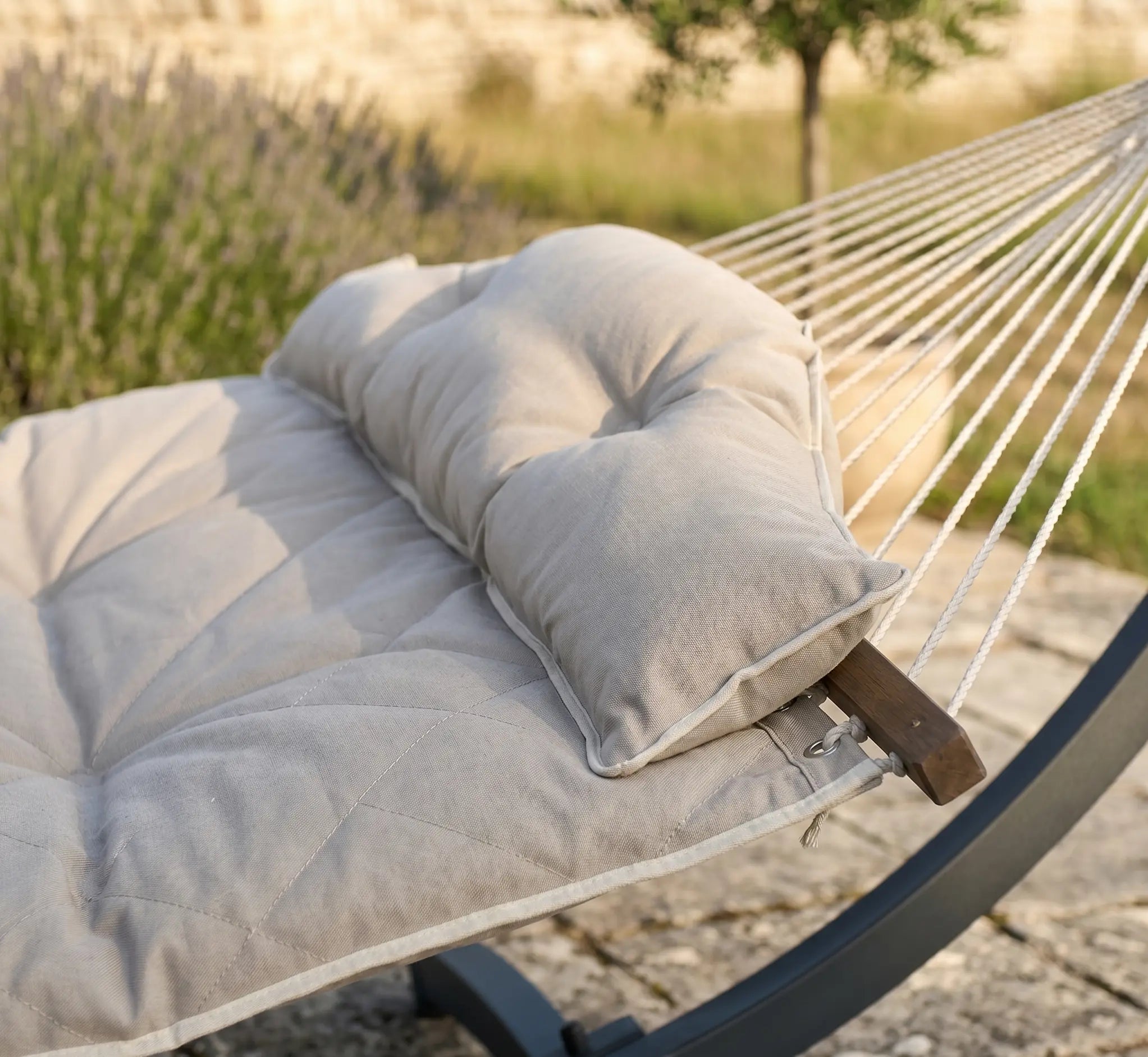 Zoom sur un hamac rembourré beige crème avec oreiller capitonné, montrant les coutures géométriques du matelas et la barre d'écartement en bois sombre sur une terrasse ensoleillée.