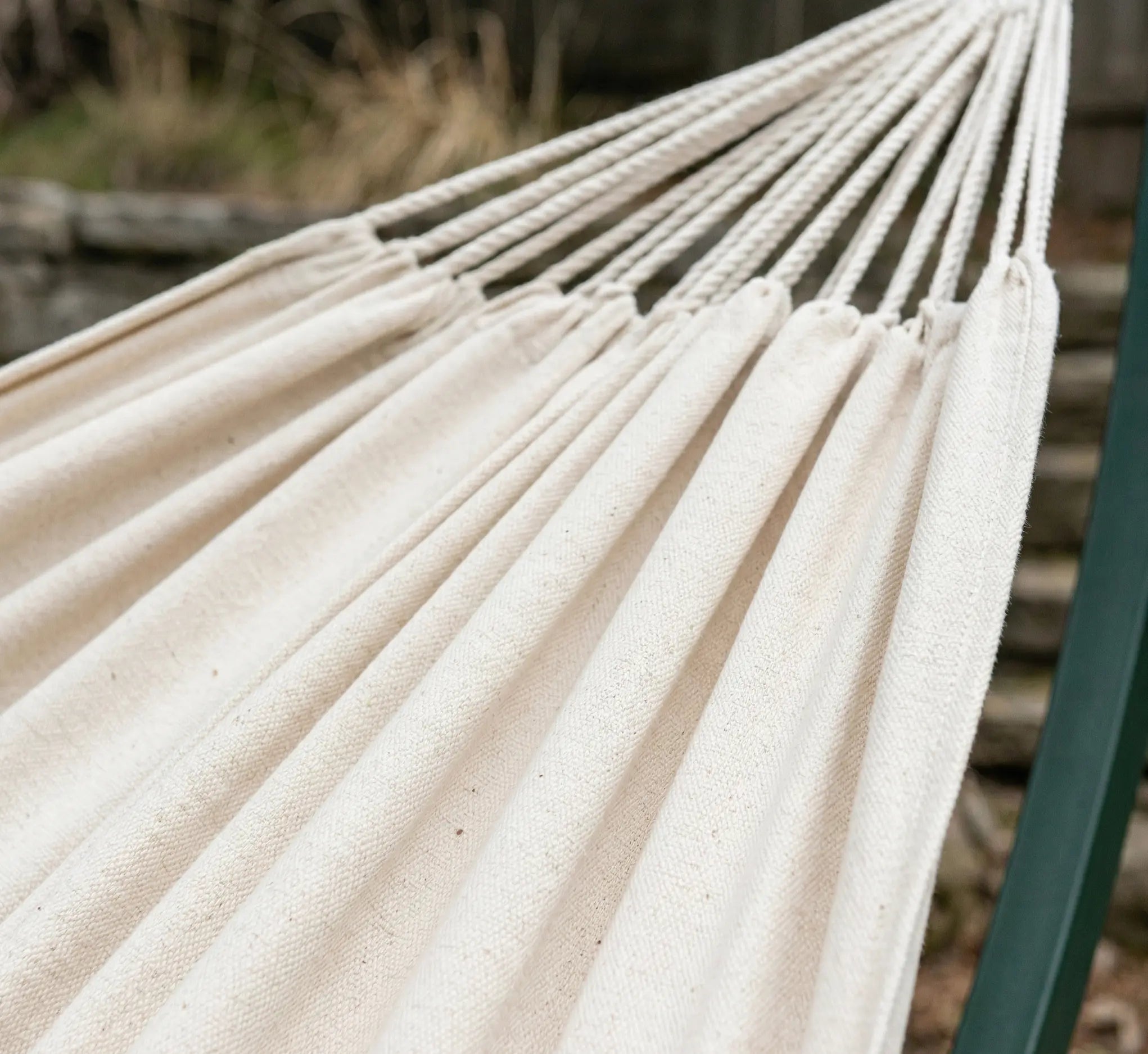 Zoom sur les finitions d'une toile de hamac en coton blanc crème haute densité montrant le tressage des suspentes et la qualité textile premium sur un support vert forêt