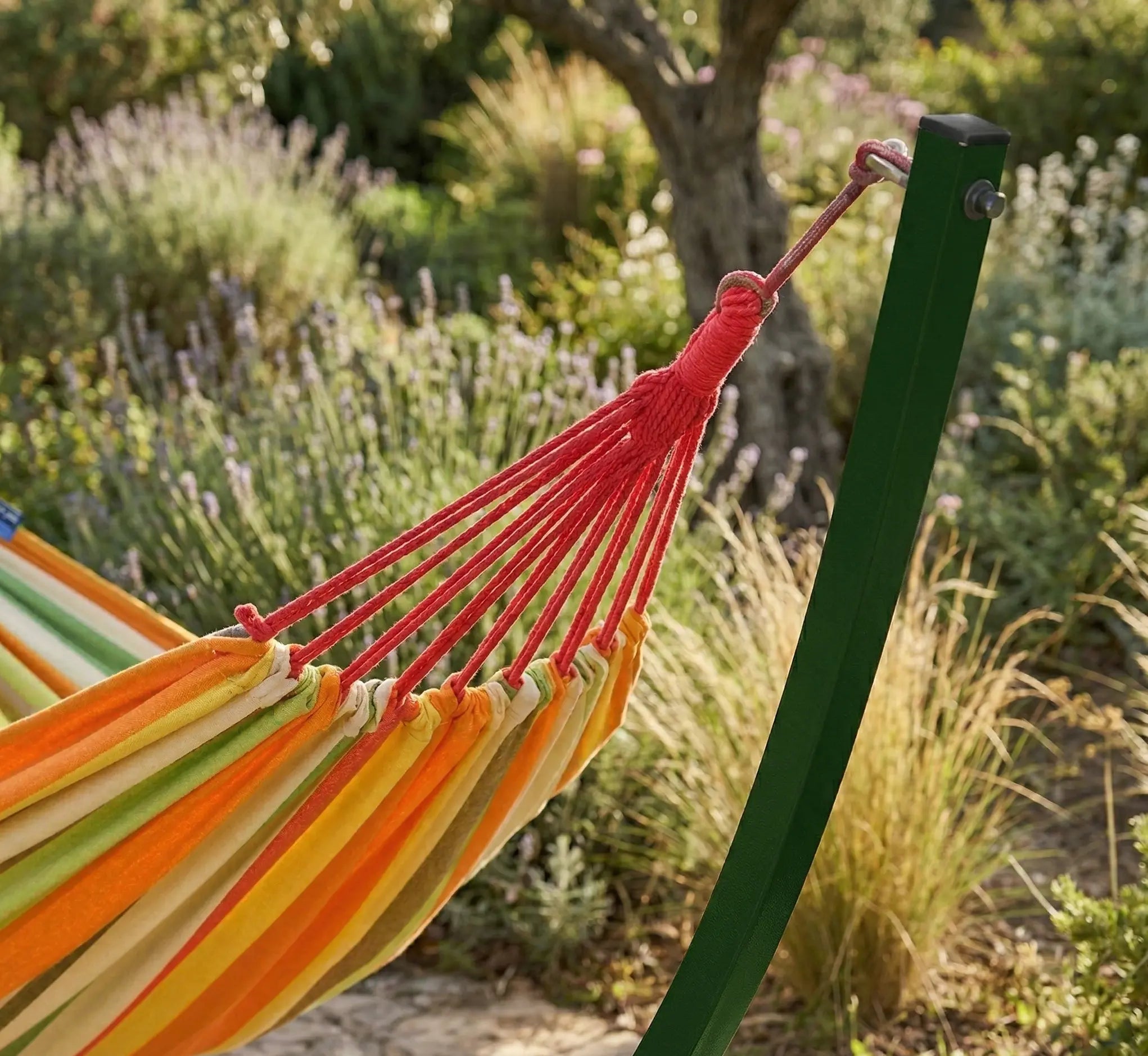 Détail du nœud de suspension renforcé et des cordages rouges d'un hamac orange sunset fixé sur un montant en acier vert forêt dans un jardin méditerranéen