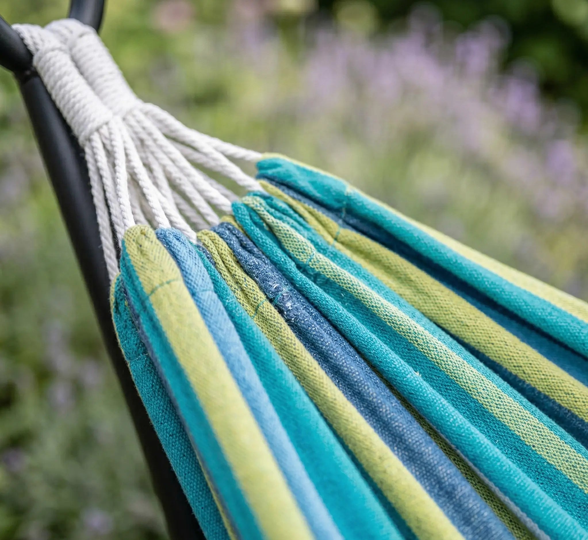 Gros plan sur les cordes de suspension en coton blanc et le maillage serré d'un hamac de luxe aux rayures vert lime et bleu turquoise dans un jardin fleuri