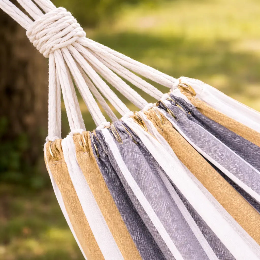 Gros plan sur les œillets métalliques dorés et les cordes de suspension blanches d'un hamac aux rayures gris, jaune moutarde et blanc.