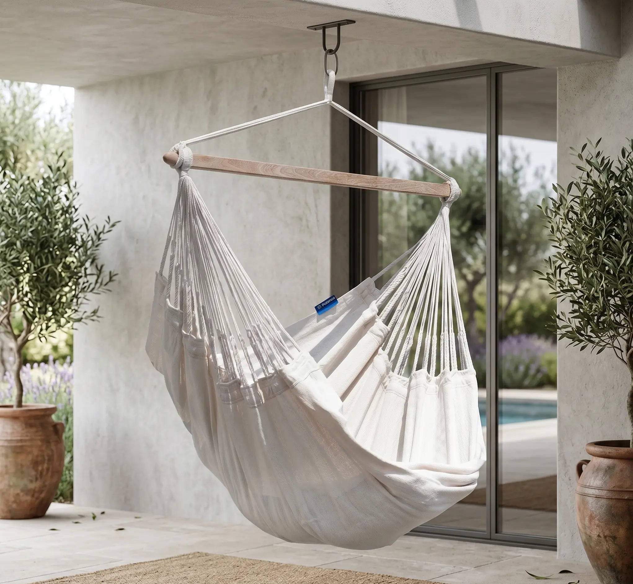 Fauteuil suspendu blanc crème avec barre en bois, fixé par un crochet métallique robuste au plafond d'une terrasse de villa moderne avec oliviers en pot et piscine en arrière-plan.
