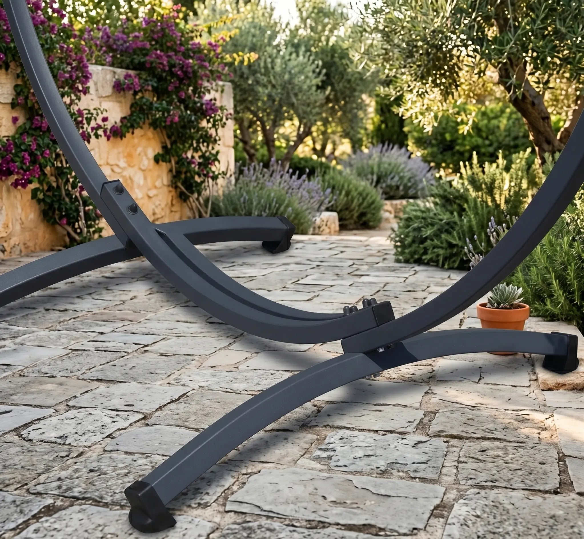 Hamac double autoportant avec toile à rayures vives (rouge, orange, jaune, violet) sur un support métallique noir incurvé, installé sur une terrasse en pierre naturelle entourée de lavande et d'un muret en terre cuite.