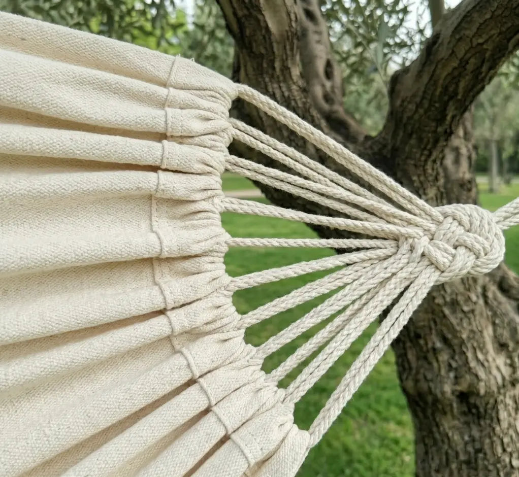 Ambiance bohème avec un hamac brésilien en coton écru naturel suspendu entre deux arbres dans un grand jardin ensoleillé, avec vue sur la campagne.