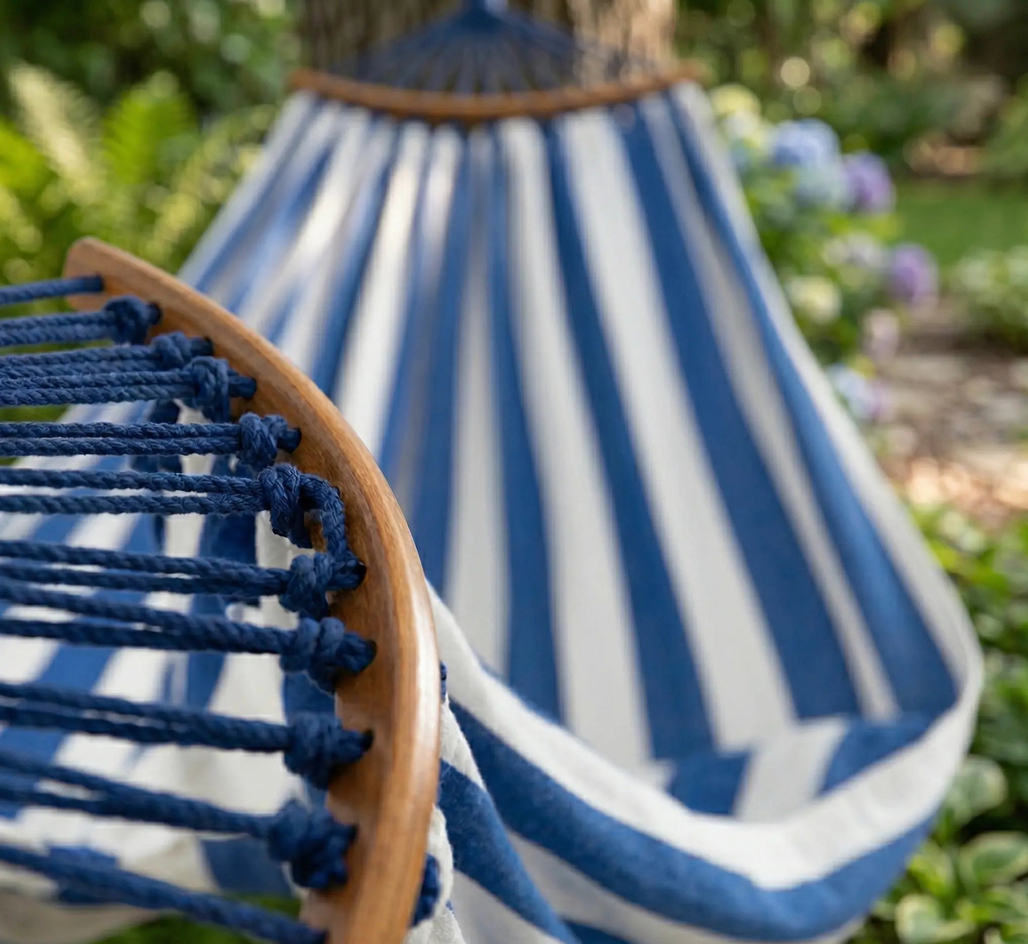 Vue d'ambiance d'un hamac de jardin à rayures bleu et blanc, avec barres d'écartement en bois et cordes de suspension bleu marine. Arrière-plan flou d'un jardin verdoyant avec hortensias.