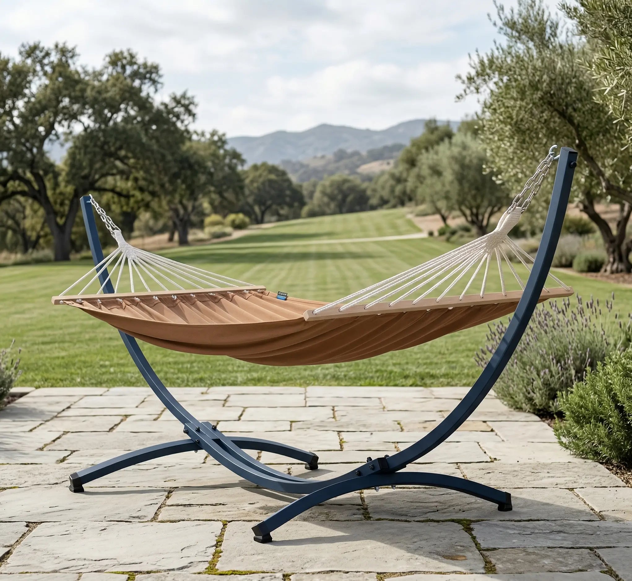 Hamac complet avec support courbé en arc anthracite et toile moka matelassée à barres bois installé sur une terrasse en dalles de pierre face à un grand jardin paysager avec oliviers