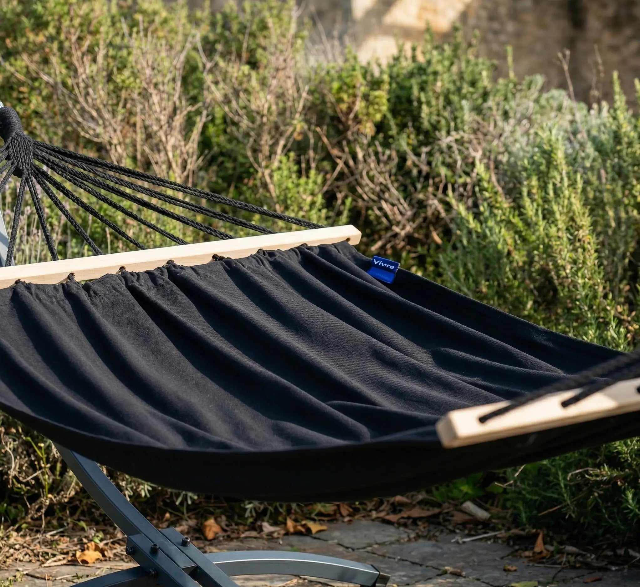 Zoom sur un hamac de luxe à barres en bois clair et toile noire premium installé sur un support anthracite robuste dans un jardin paysager