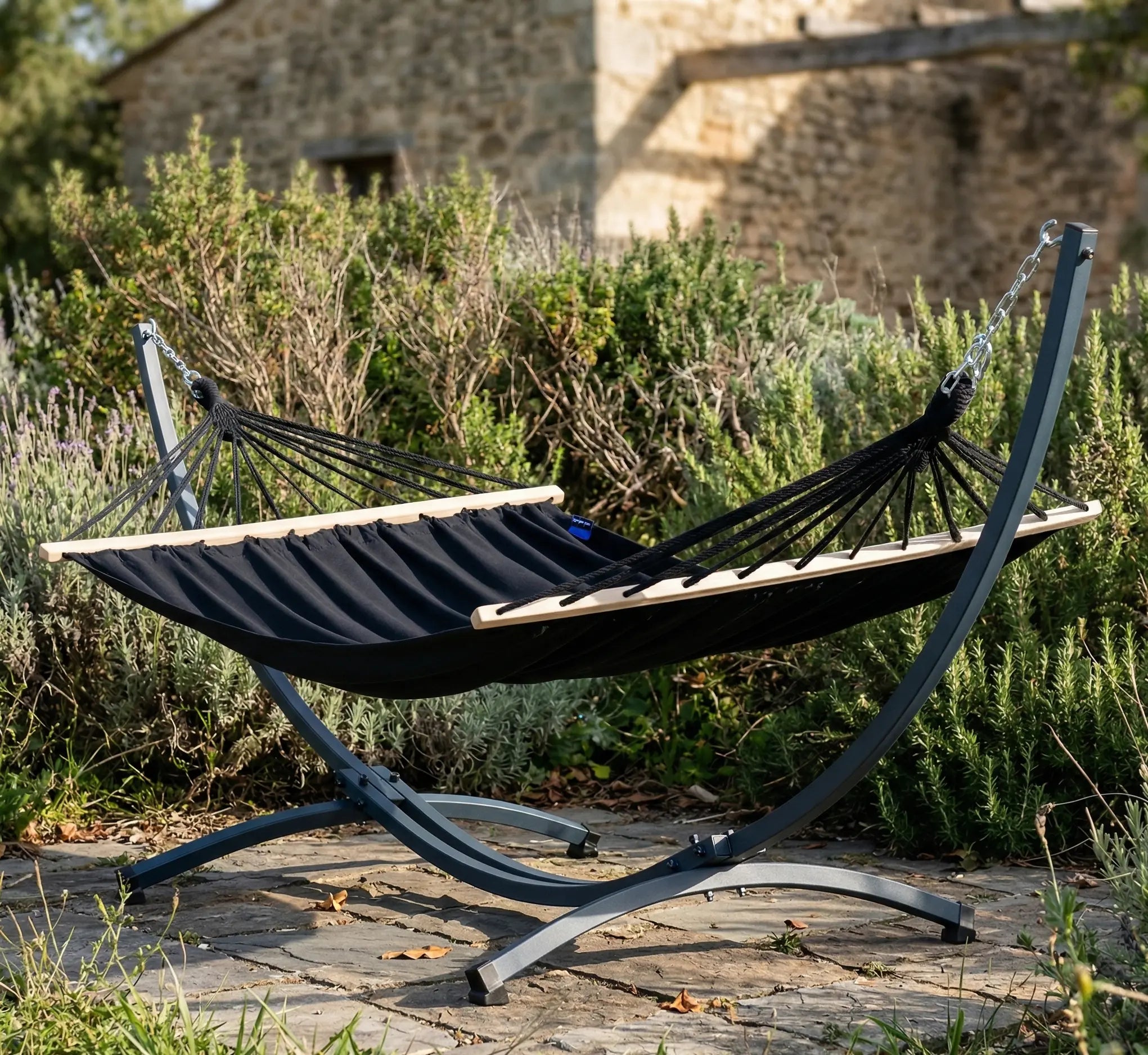 Hamac haut de gamme avec support courbé en arc anthracite et toile noire tendue par des barres en bois naturel installé sur une terrasse en dalles de pierre dans un jardin paysager ombragé
