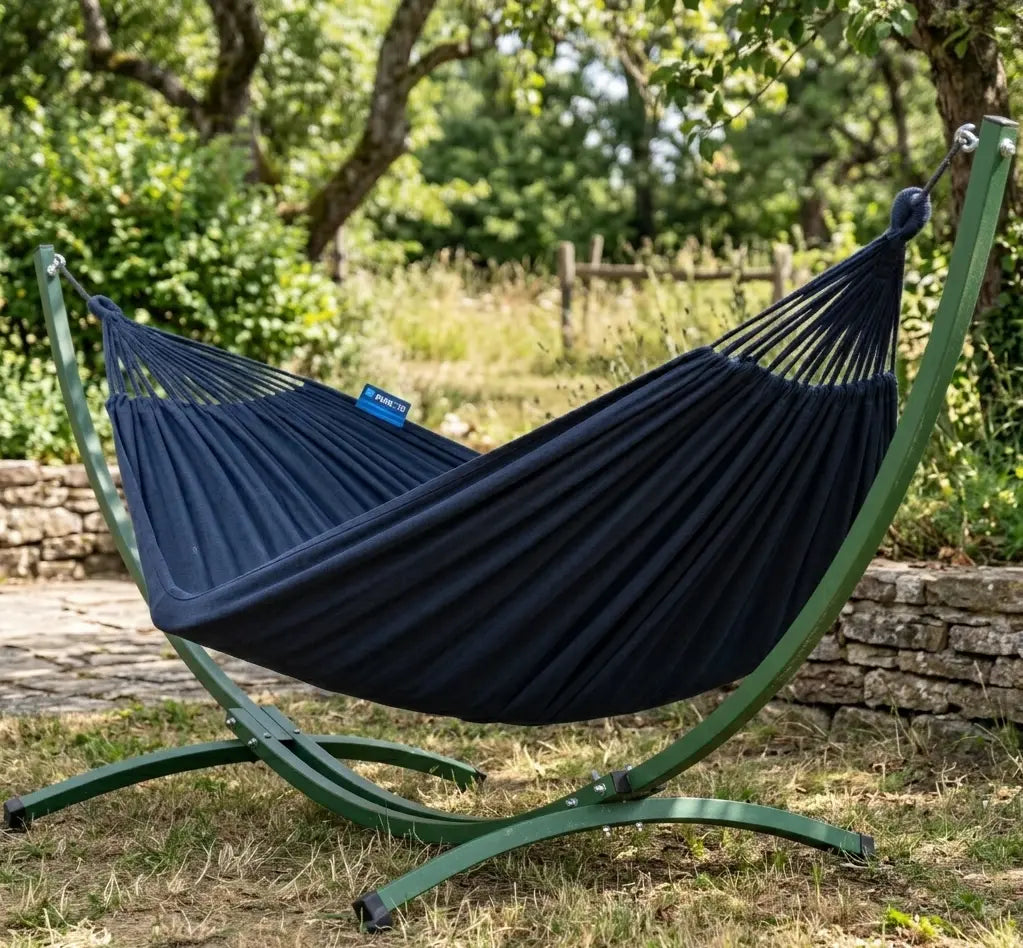 Hamac autoportant avec support courbé design en acier vert forêt et toile en coton bleu marine installé dans un jardin arboré avec muret en pierre sèche