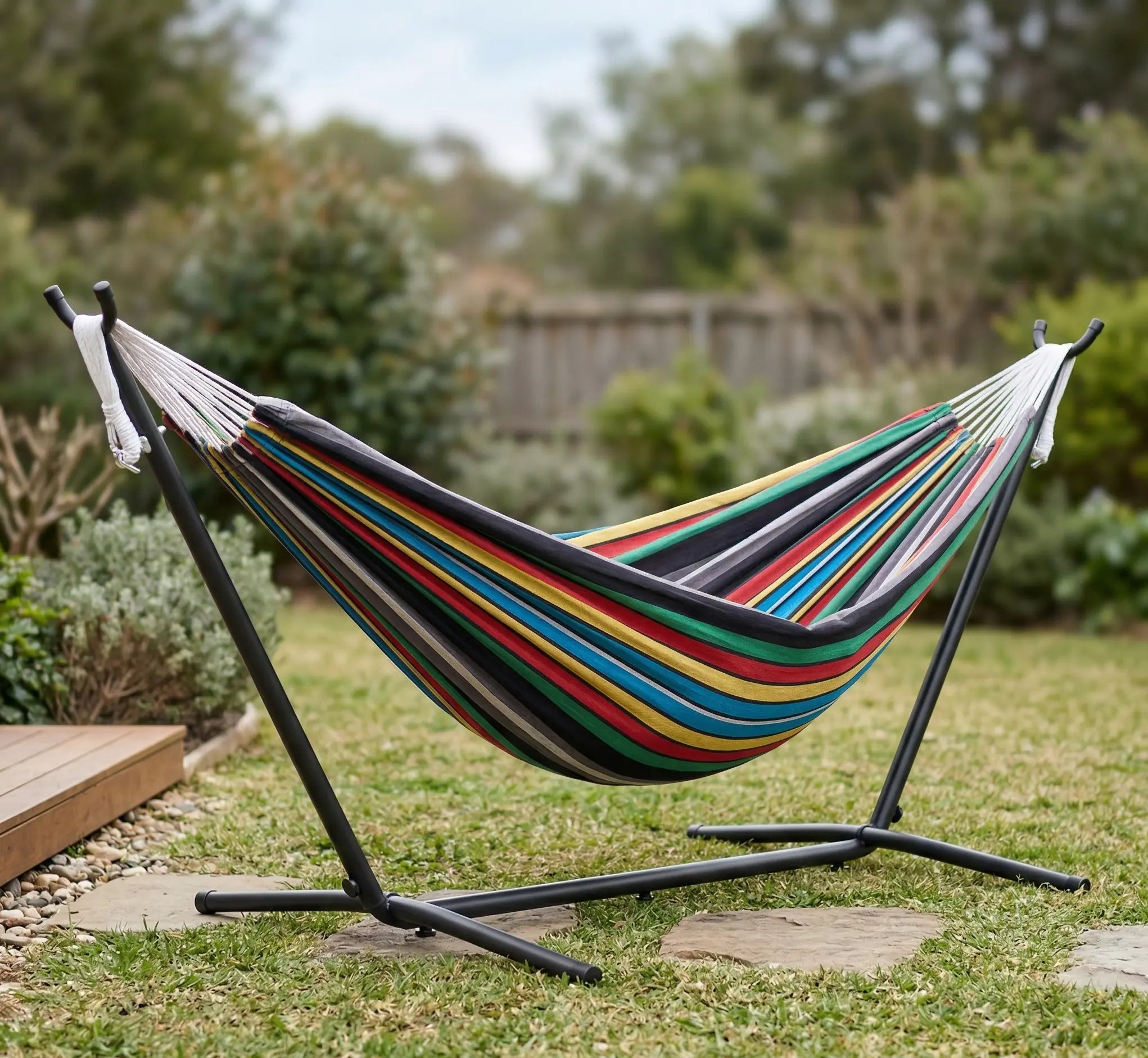 Hamac autoportant complet avec support en acier noir installé sur une pelouse dans un jardin paysager montrant une toile en coton aux rayures vives vert jaune rouge et noir