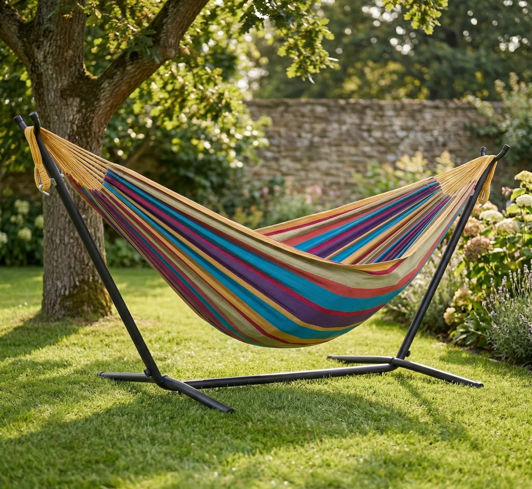 Hamac autoportant complet avec support en métal noir installé sur une pelouse verte dans un jardin ensoleillé avec toile en coton aux rayures chaudes ocre jaune violet et turquoise