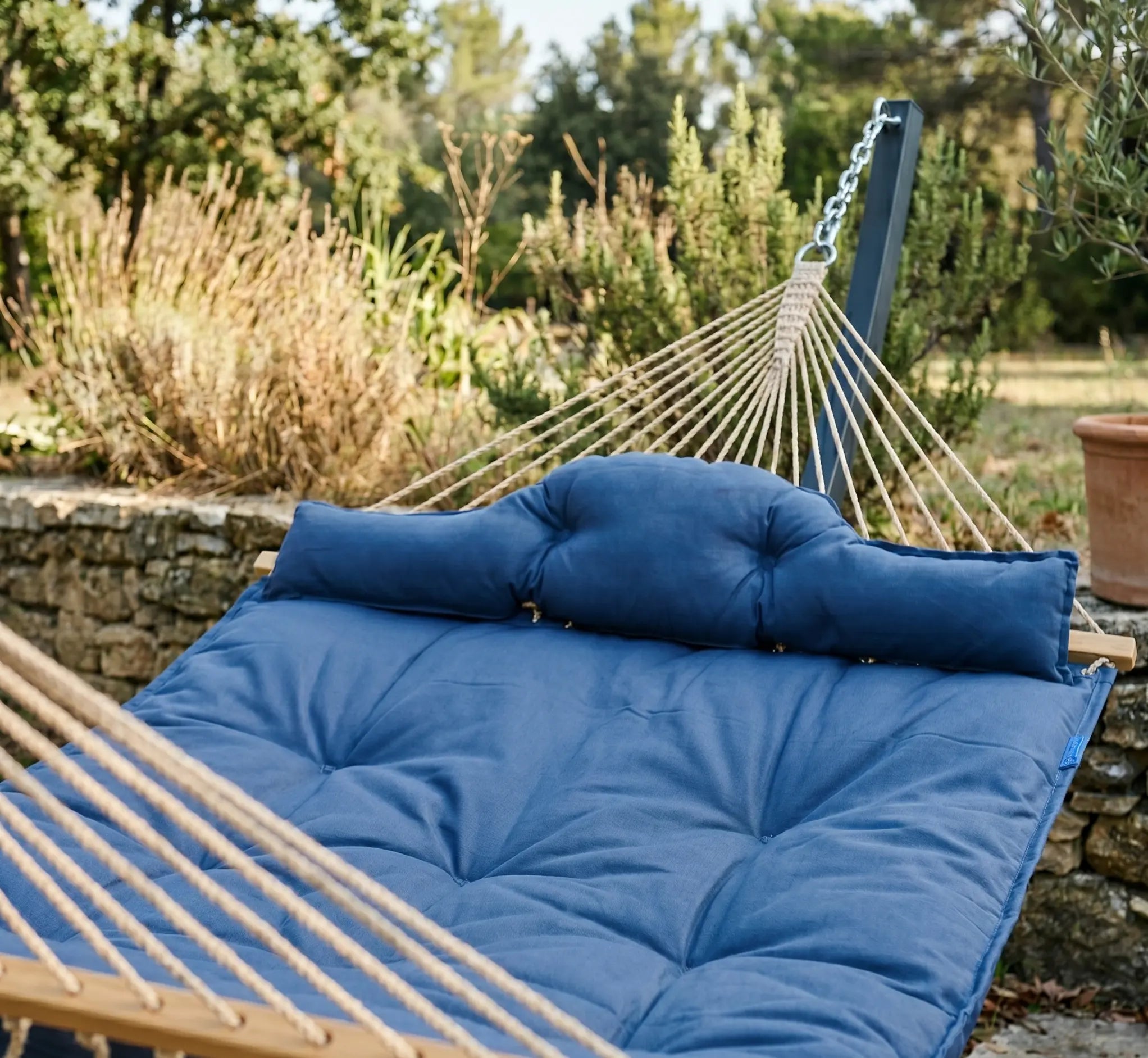 Vue rapprochée d'un lit hamac rembourré bleu marine avec oreiller capitonné intégré, suspendu par des chaînes en acier et des cordes robustes dans un jardin avec muret en pierre et oliviers.